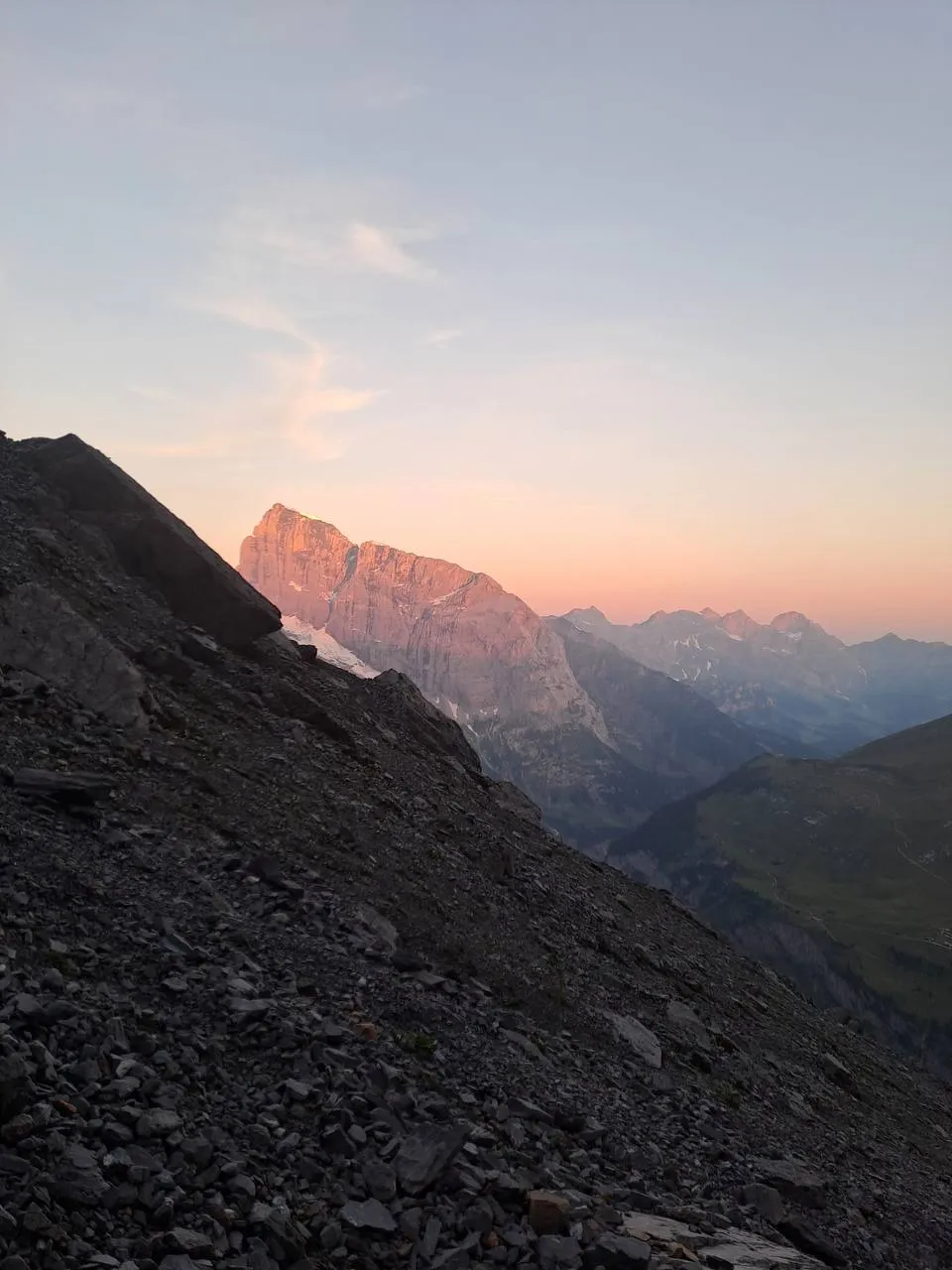 The Schlossberg south wall glowing pink in the alpenglow, with a dark scree slope in front and ranks of peaks in the morning haze.