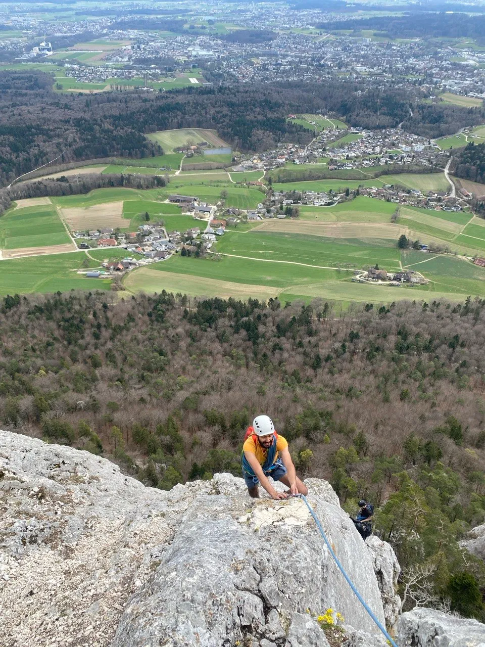 A climber holds onto the rock face while belaying, with villages and farmland far below in the Jura.