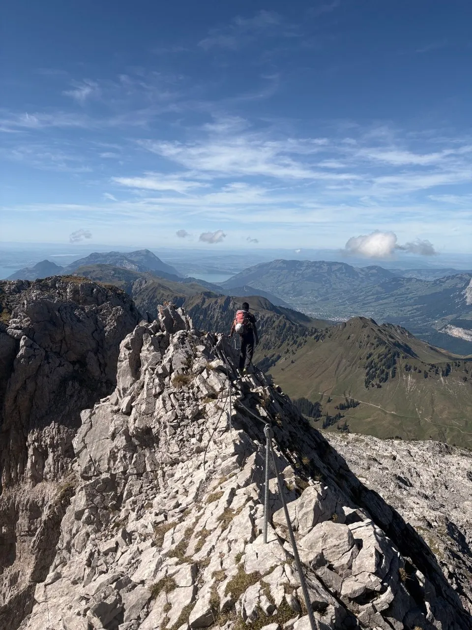 Excursionista en una cresta rocosa expuesta y asegurada con cadenas, con un lago encajonado y un amplio panorama de montañas al fondo.