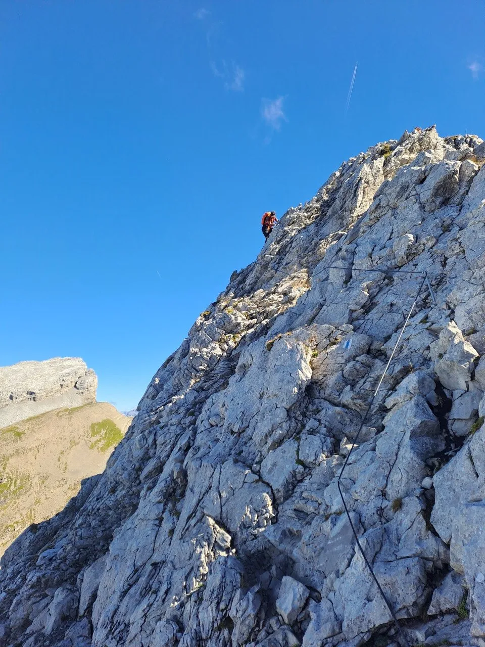 A climber in orange near the upper edge of the grey limestone wall, a cable in the rock, a neighbouring peak to the left under a blue sky.