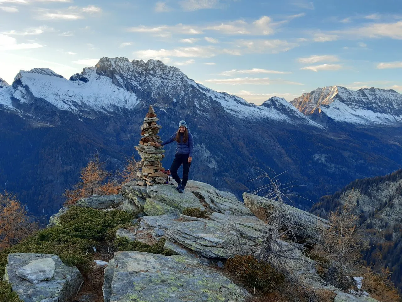 A hiker beside a tall dry-stone cairn on a rocky ridge, golden autumn larches falling away to the valley and snow-dusted peaks behind.