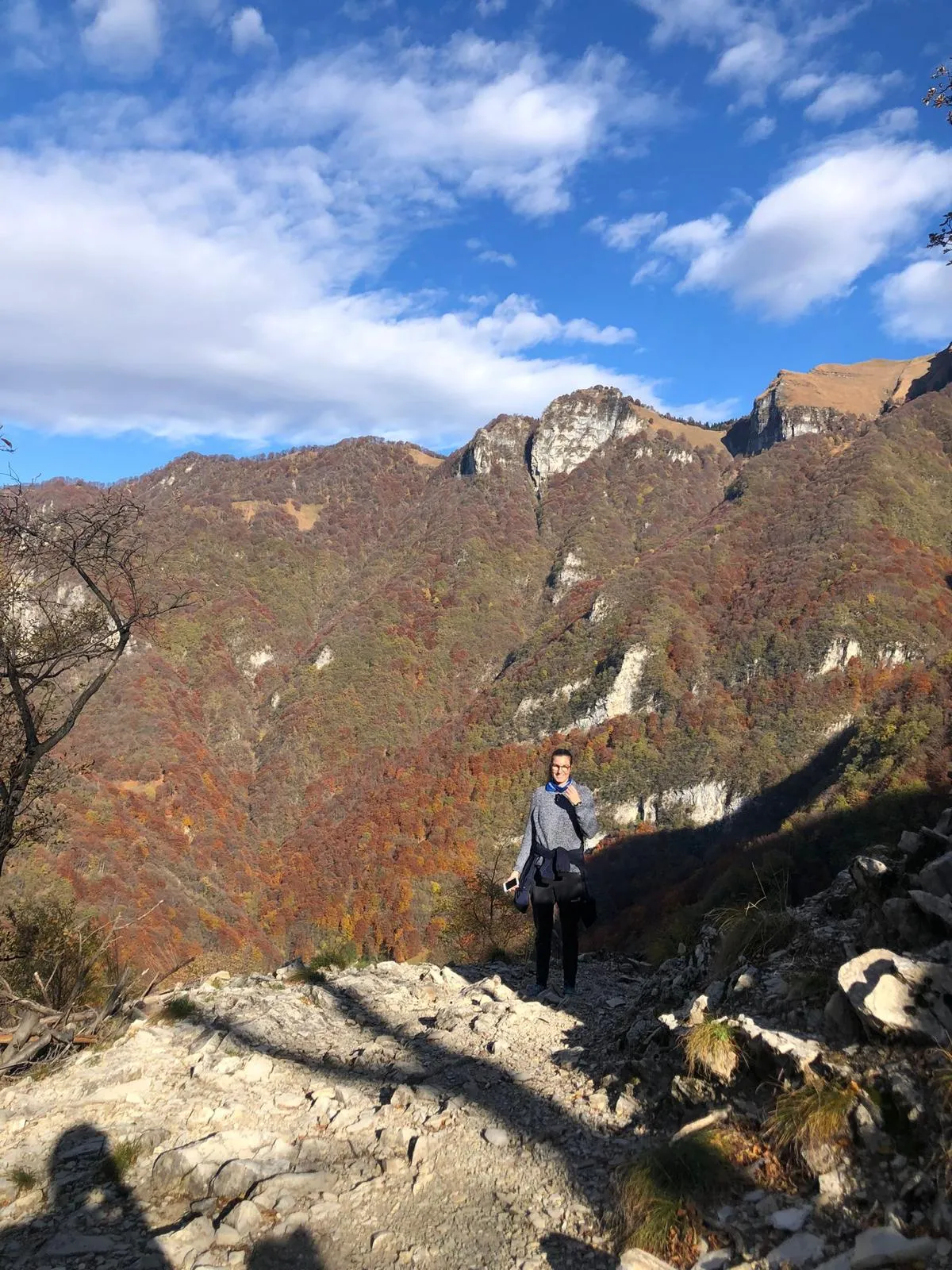 A hiker on a stony trail with autumn-red forested slopes and craggy peaks behind.