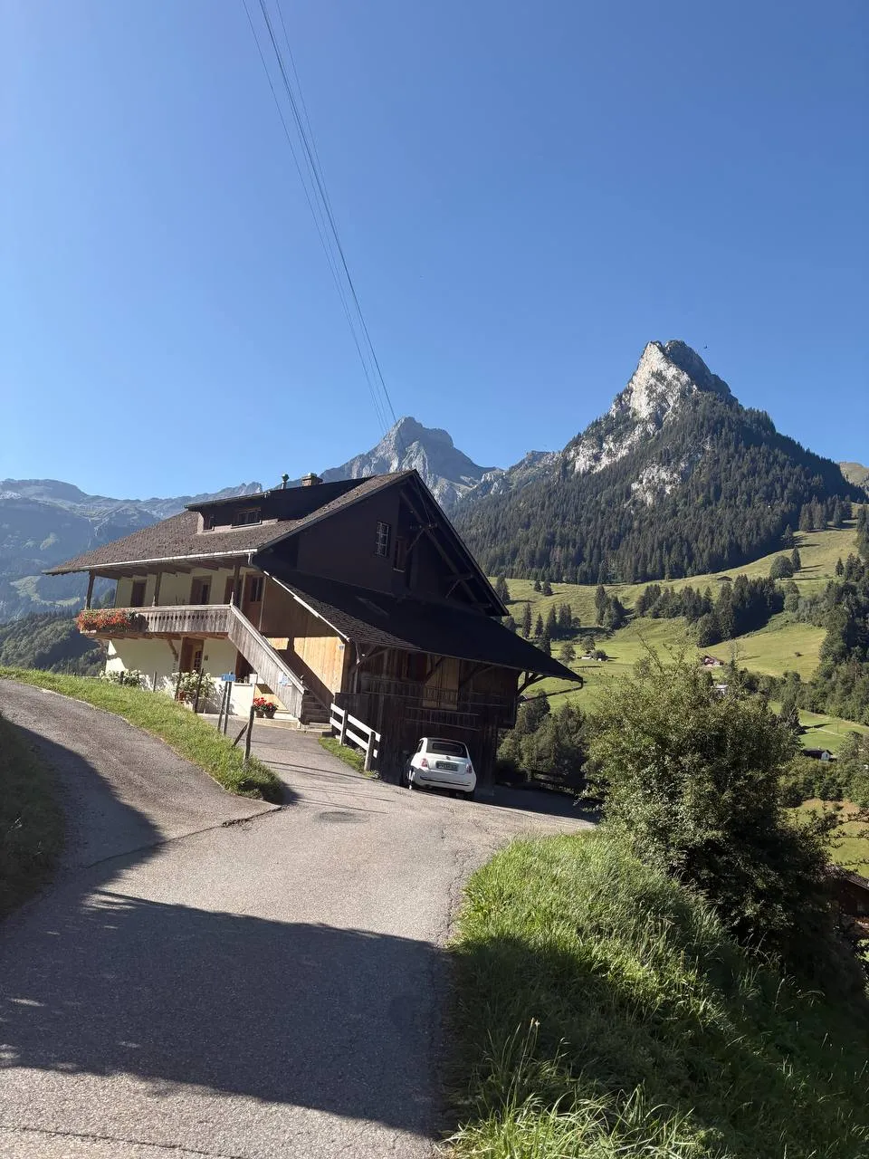 A big Bernese farmhouse with a wooden balcony beside the road, a pointed rock peak behind it in the morning light.