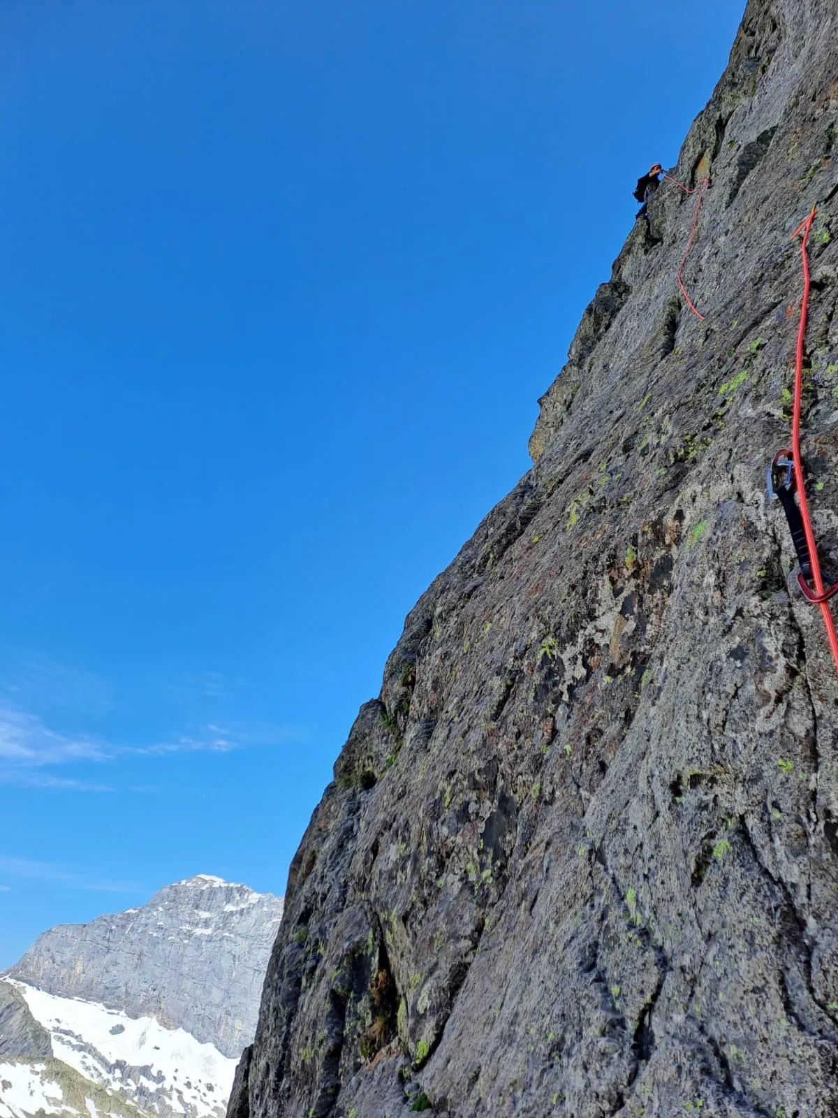 A lead climber high on the steep grey wall of the Gwächten, a red rope in the rock, blue sky and a snow-capped peak beyond.