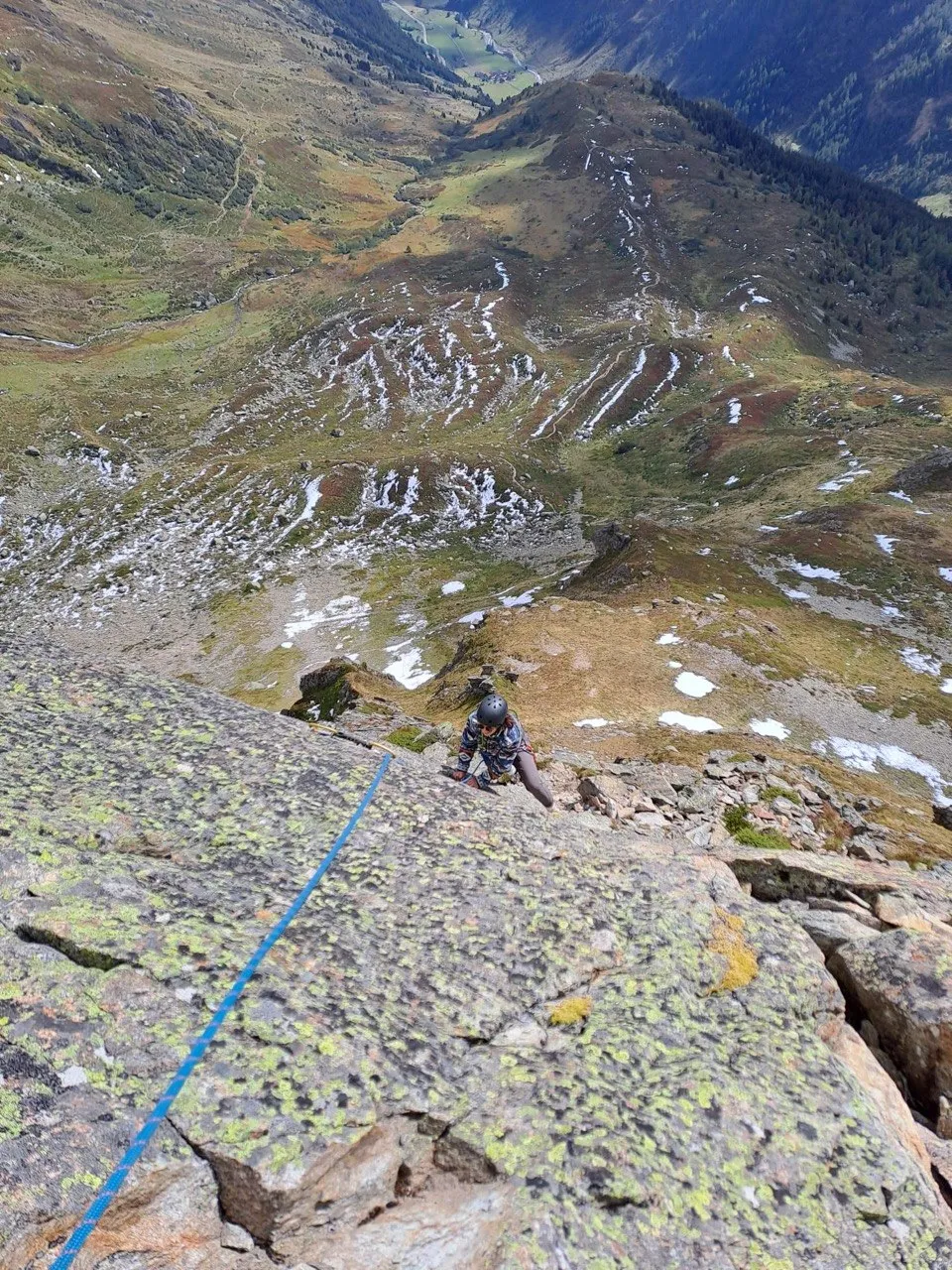 A roped climber on a lichen-covered slab, a snow-patched valley far below.
