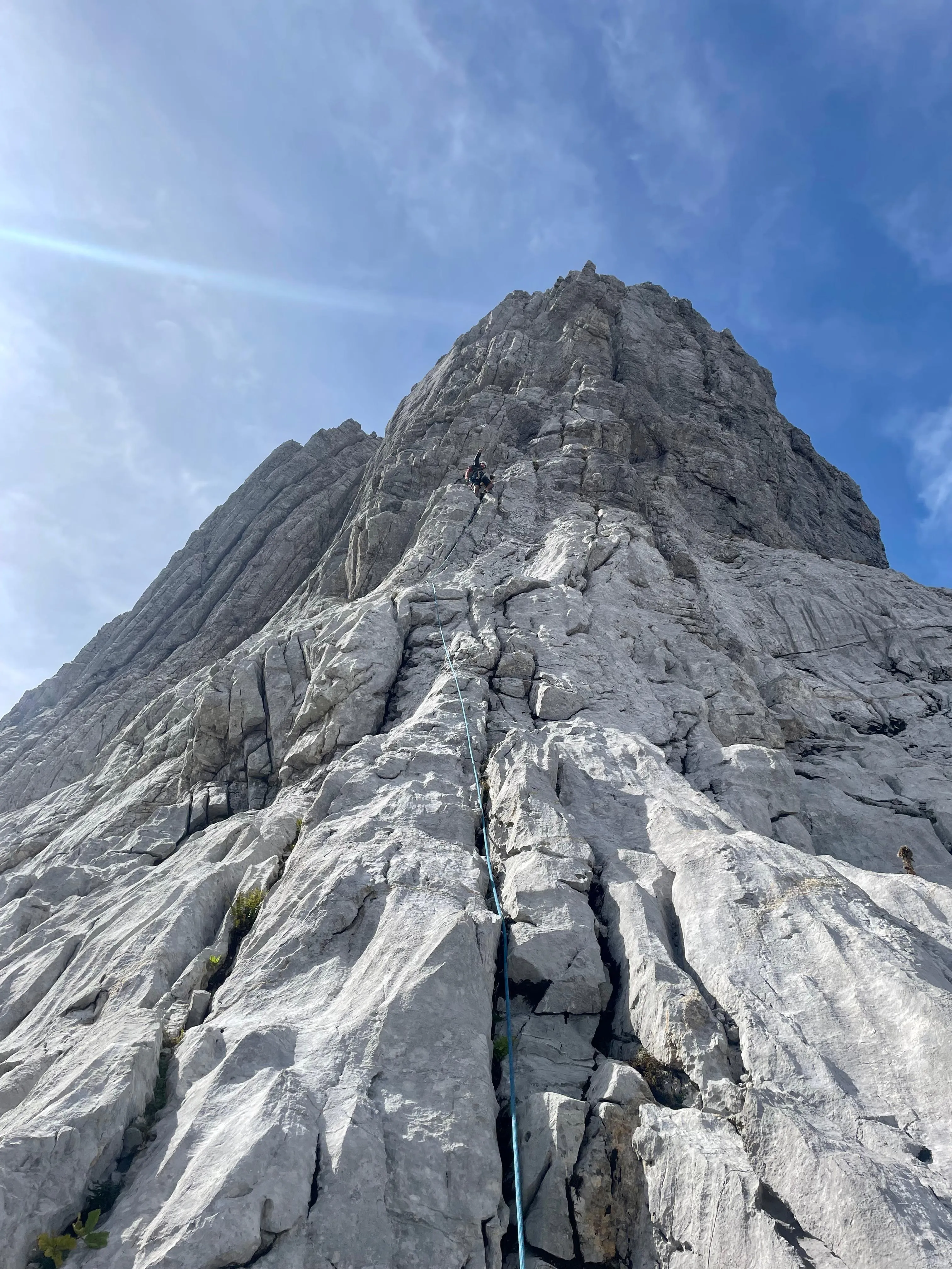 Climber on the east ridge below the towering Altmann summit pyramid, the blue rope running down the grey limestone slab against a blue sky.