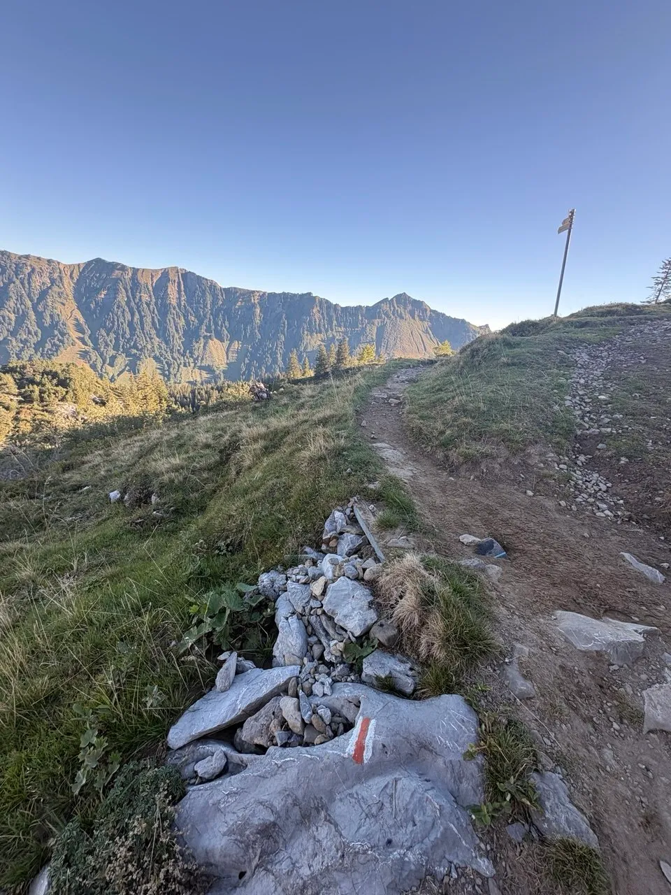 Narrow mountain path with a red-and-white waymark painted on rock, a sunlit ridge beyond.