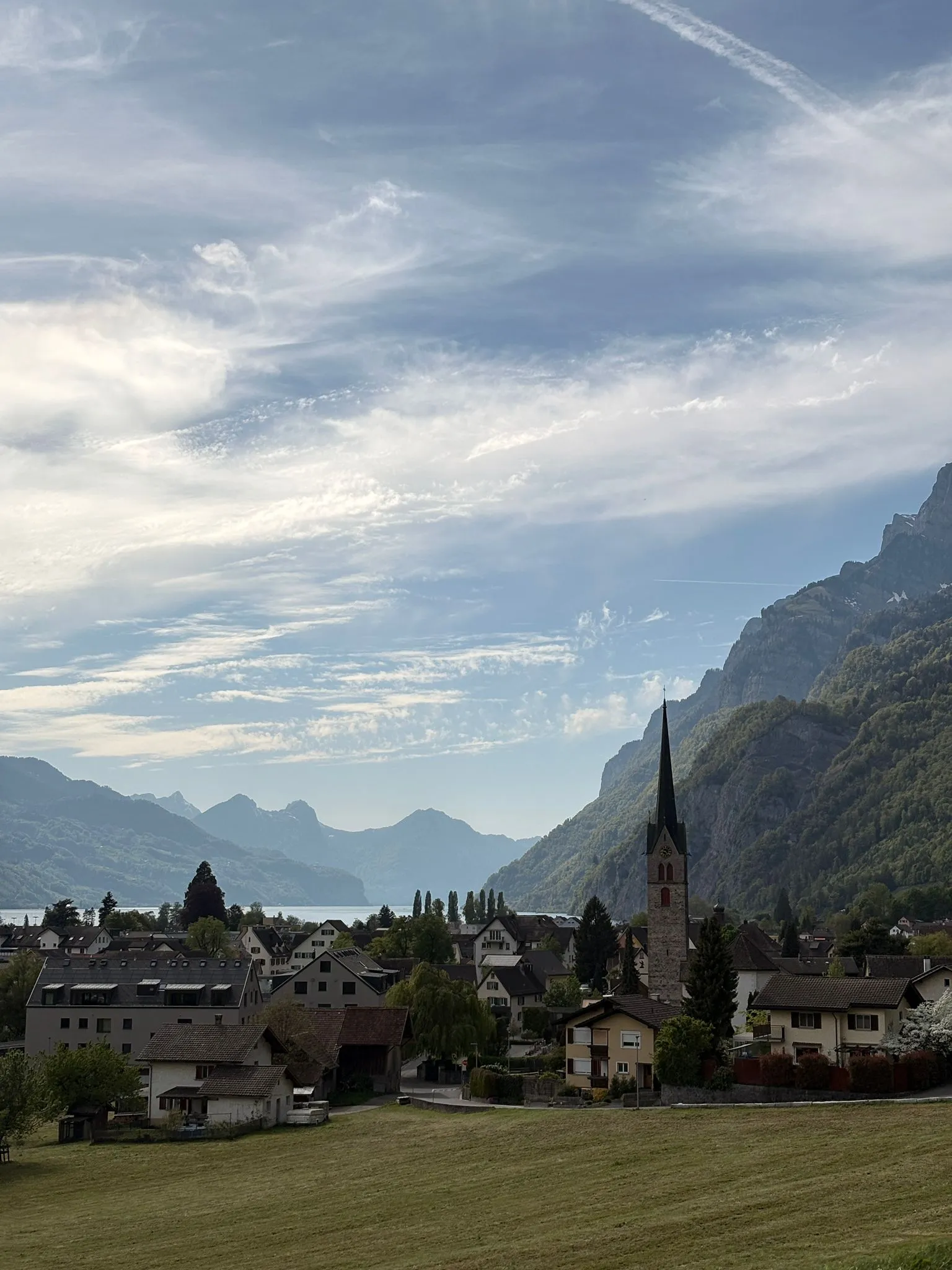 A village church spire below the Churfirsten in soft early-morning light, with the Walensee in the distance.