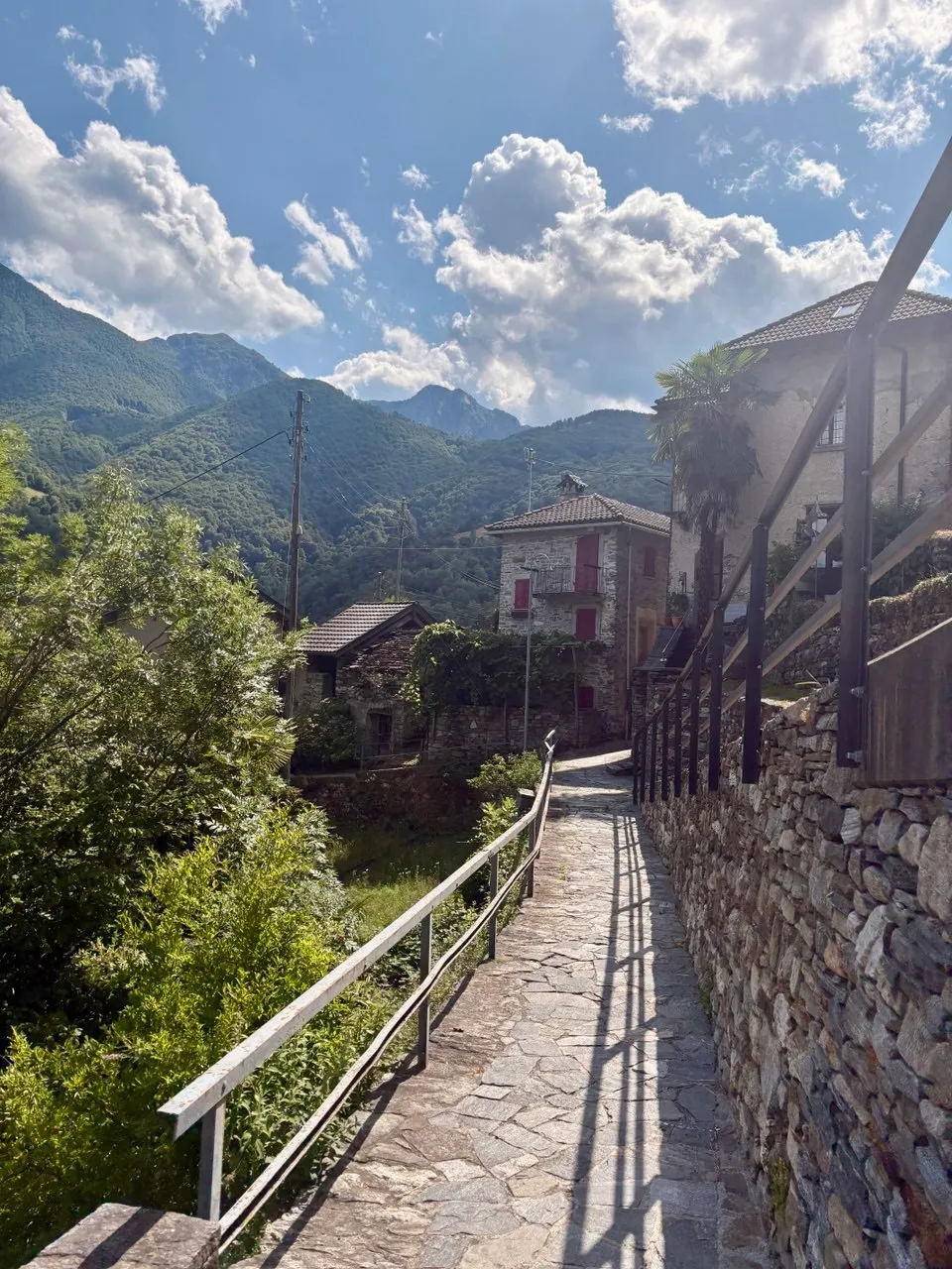 A paved stone path with a railing leading down through a Ticino village of stone houses with red shutters, a palm tree and green mountains behind.