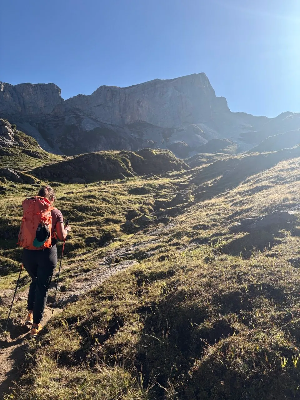 Hiker with an orange pack climbs grassy slopes in the morning light toward the rocky bulk of the Chaiserstock.
