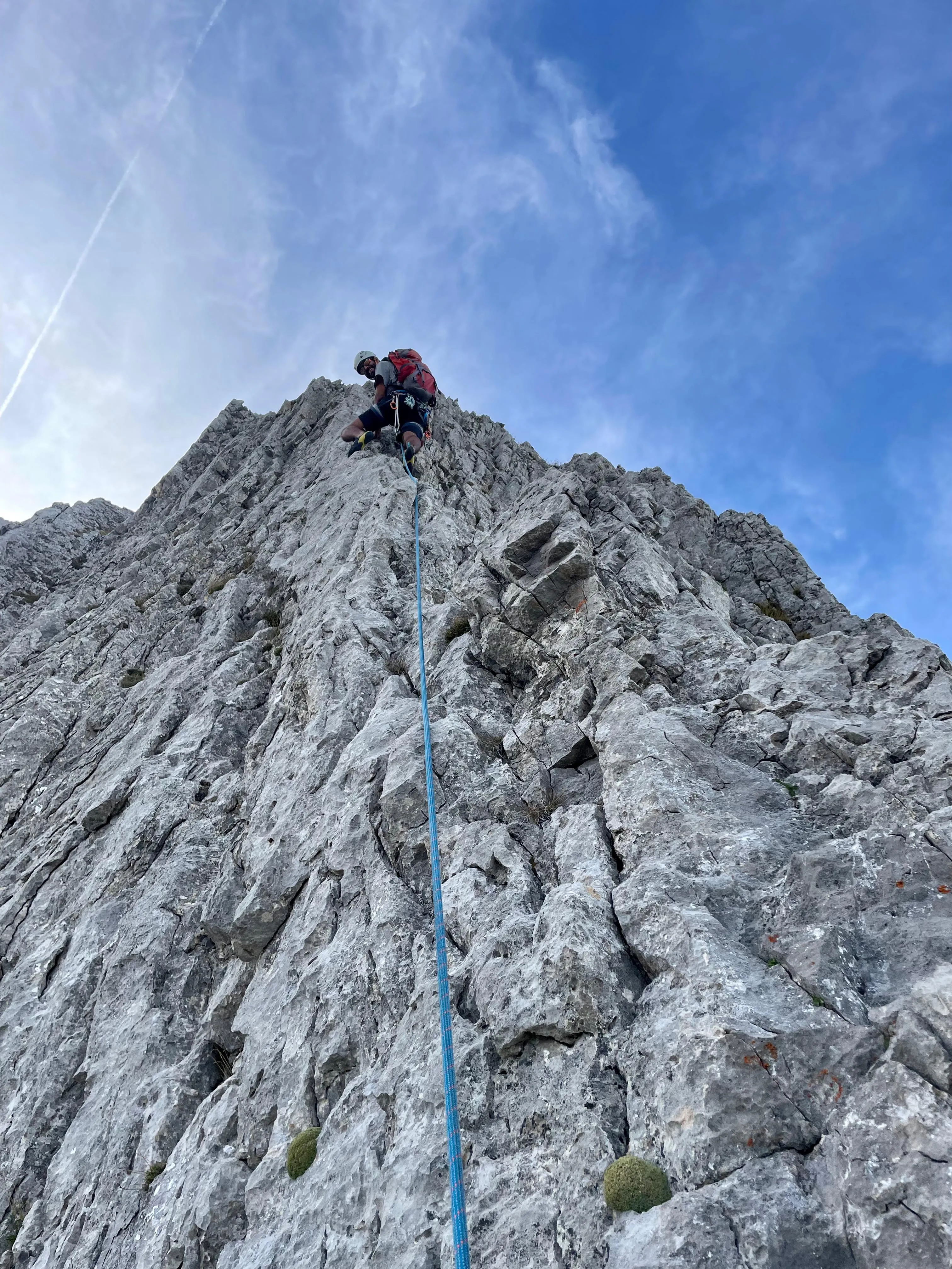 Climber with a red backpack and white helmet high on the grey ridge, the blue rope hanging into the depth beneath a blue sky.