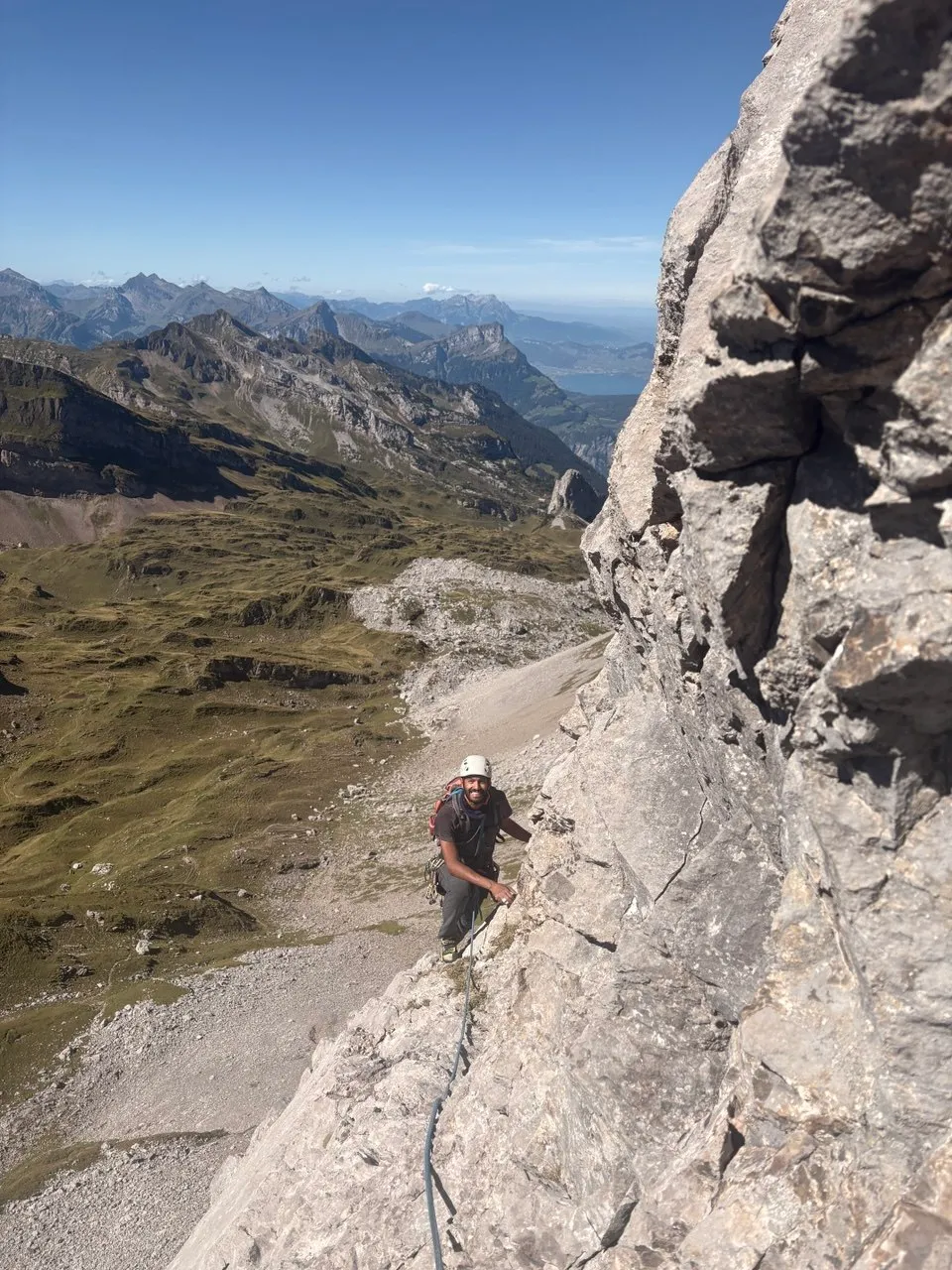A smiling climber in a helmet on the grey west face of the Chaiserstock, a green valley, a lake and layered mountain ranges far below.