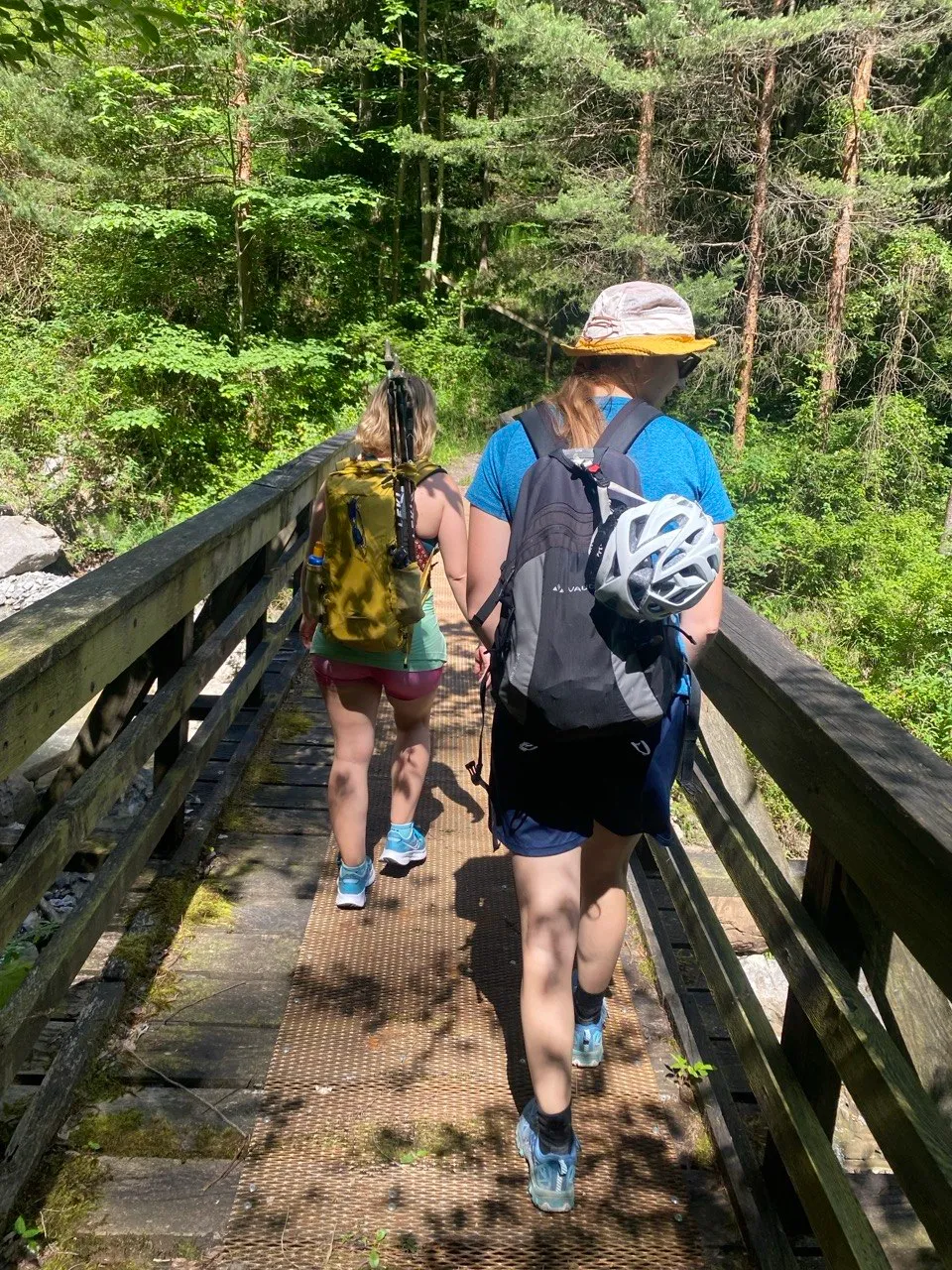 Two hikers crossing a wooden bridge on a shaded forest trail.