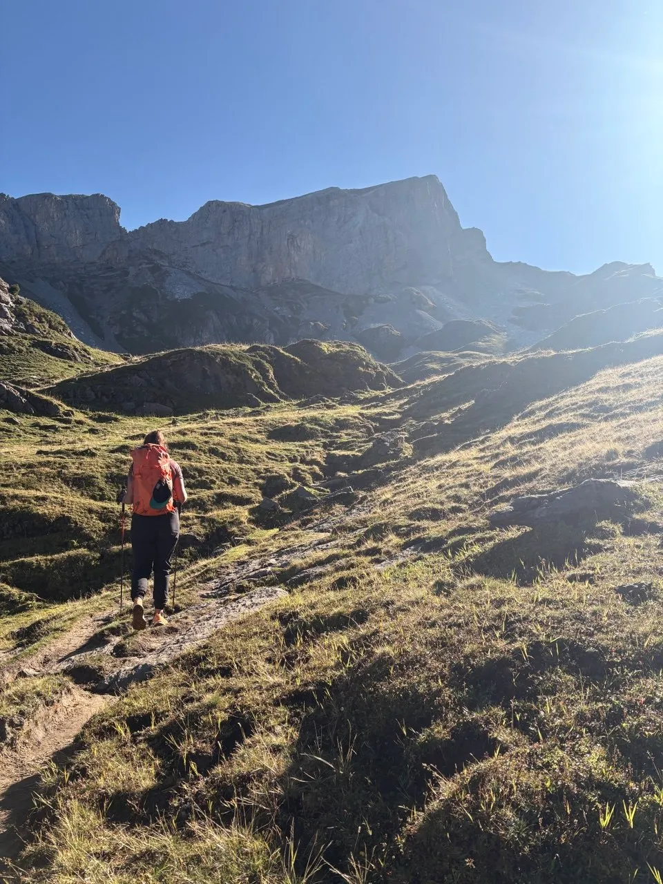 Hiker with an orange pack climbs a grassy slope below steep grey cliffs in backlight.