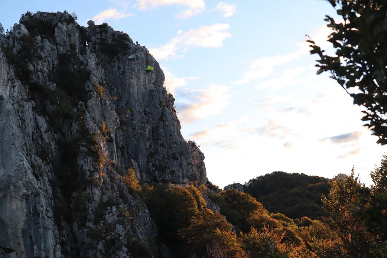 Pared de roca caliza del Sasso Grande con luz cálida del atardecer y una tienda de vivac en una repisa muy por encima del bosque.