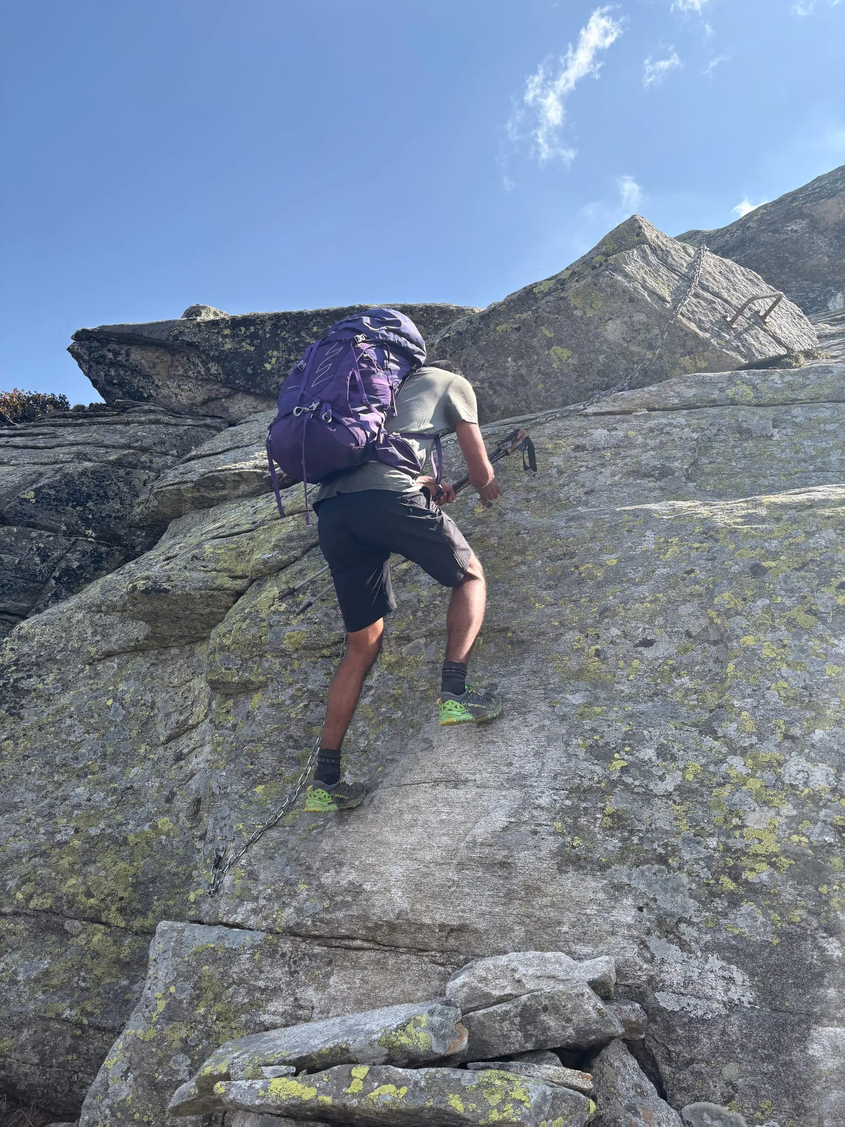 A hiker scrambles up steep granite slabs with a backpack on — the T5− crux below the Madom da Sgiof.