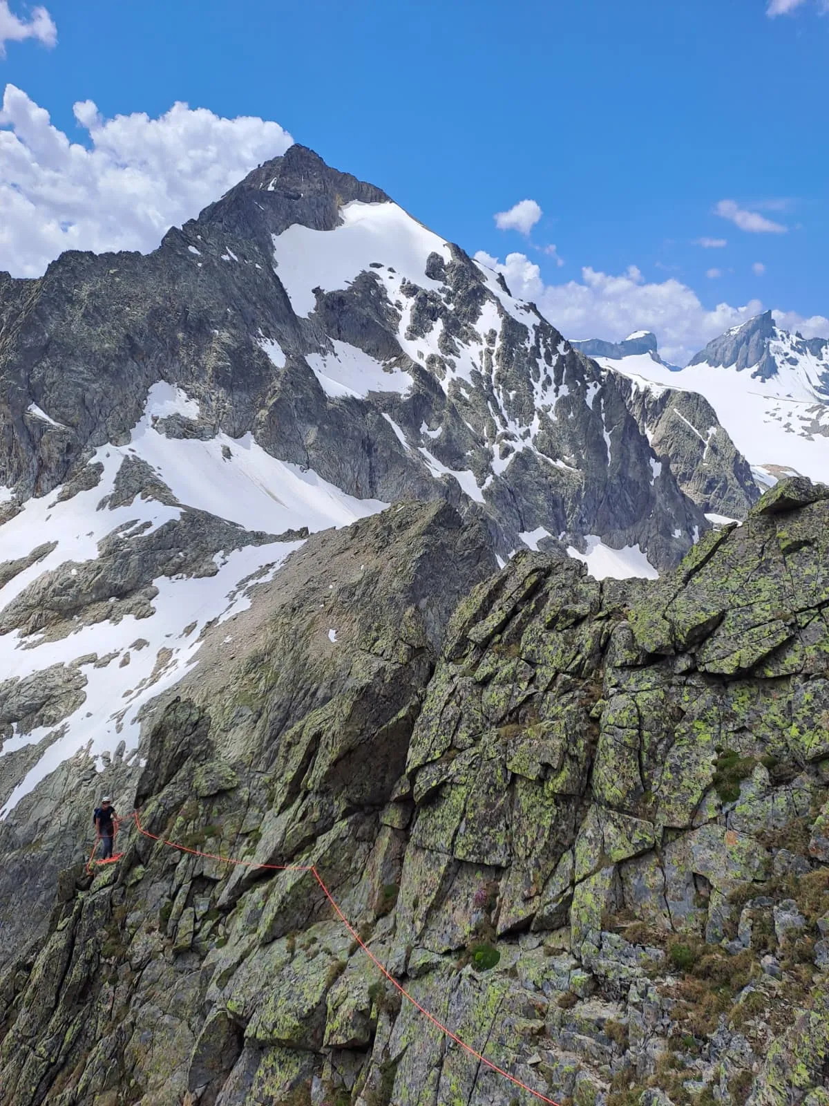 A climber on the lichen-green rock ridge, the pointed, partly snow-covered summit of the Gwächten behind under a blue sky.