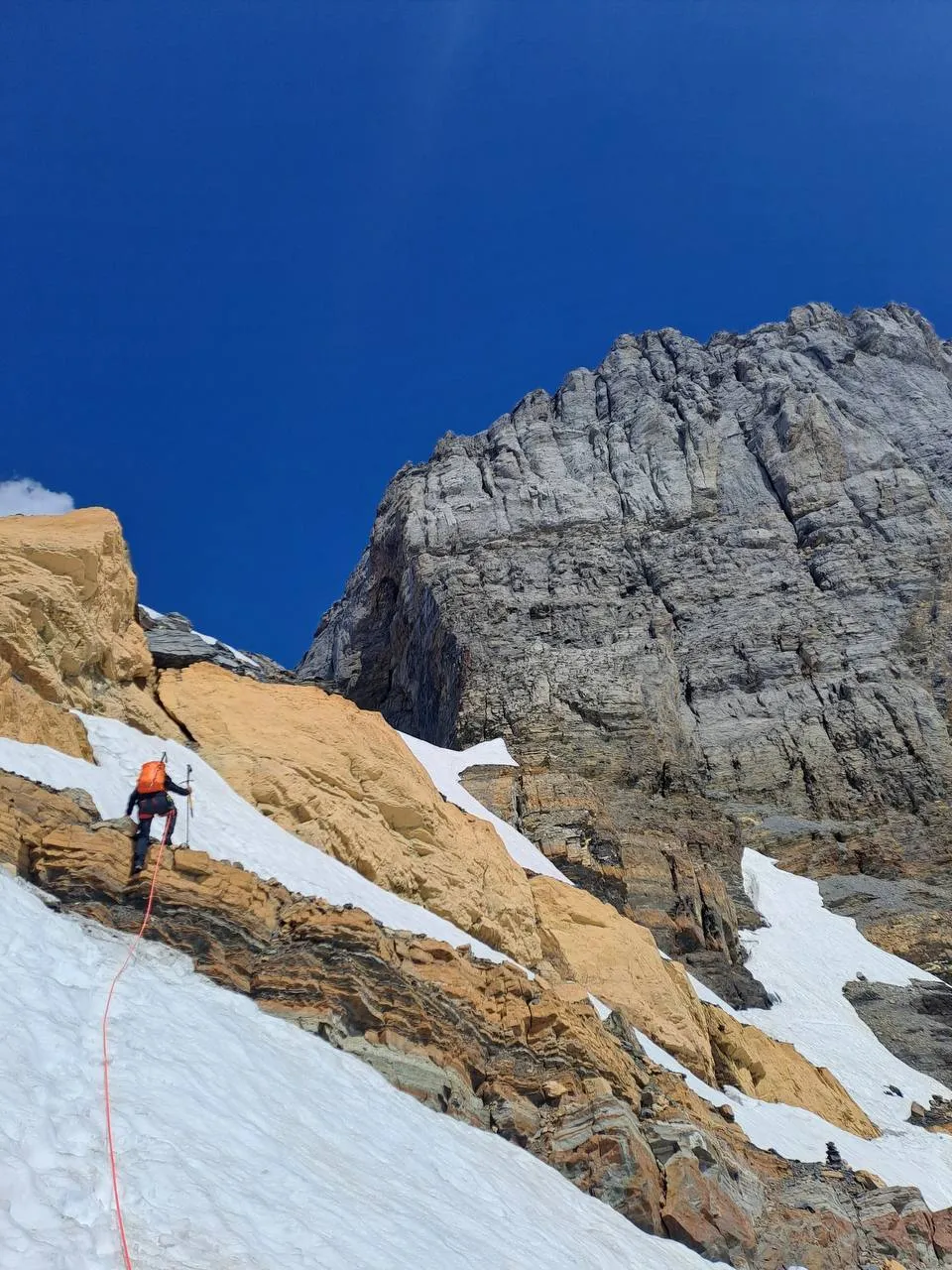 A climber in an orange helmet ascends a snowfield toward the orange-and-grey south wall of the Schlossberg, the red rope hanging below.