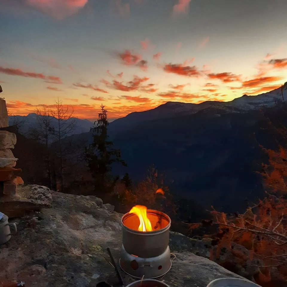 A spirit stove burning on a rock slab at dusk, autumn larches and a dry-stone cairn beside it, snow-dusted mountains under a blazing orange sunset behind.