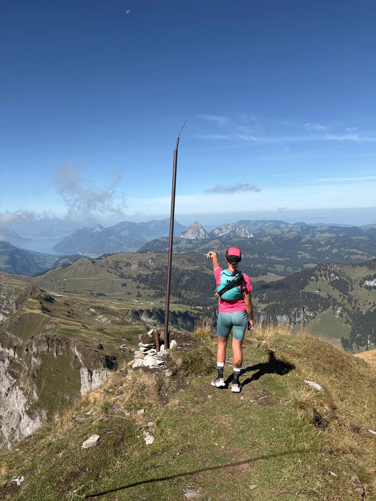 A runner stands by the iron summit pole and points across to the Mythen, Lake Lucerne below to the left and a pale moon in the sky.