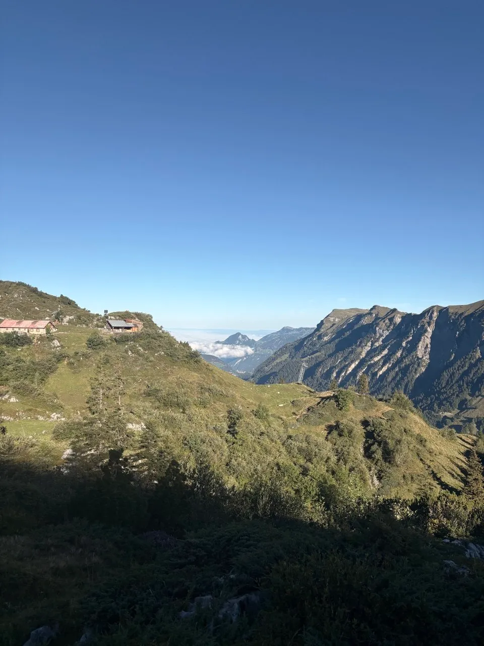 Alpine huts on a ridge with the cable car's top station, a sea of clouds over the valley and distant ranges behind.