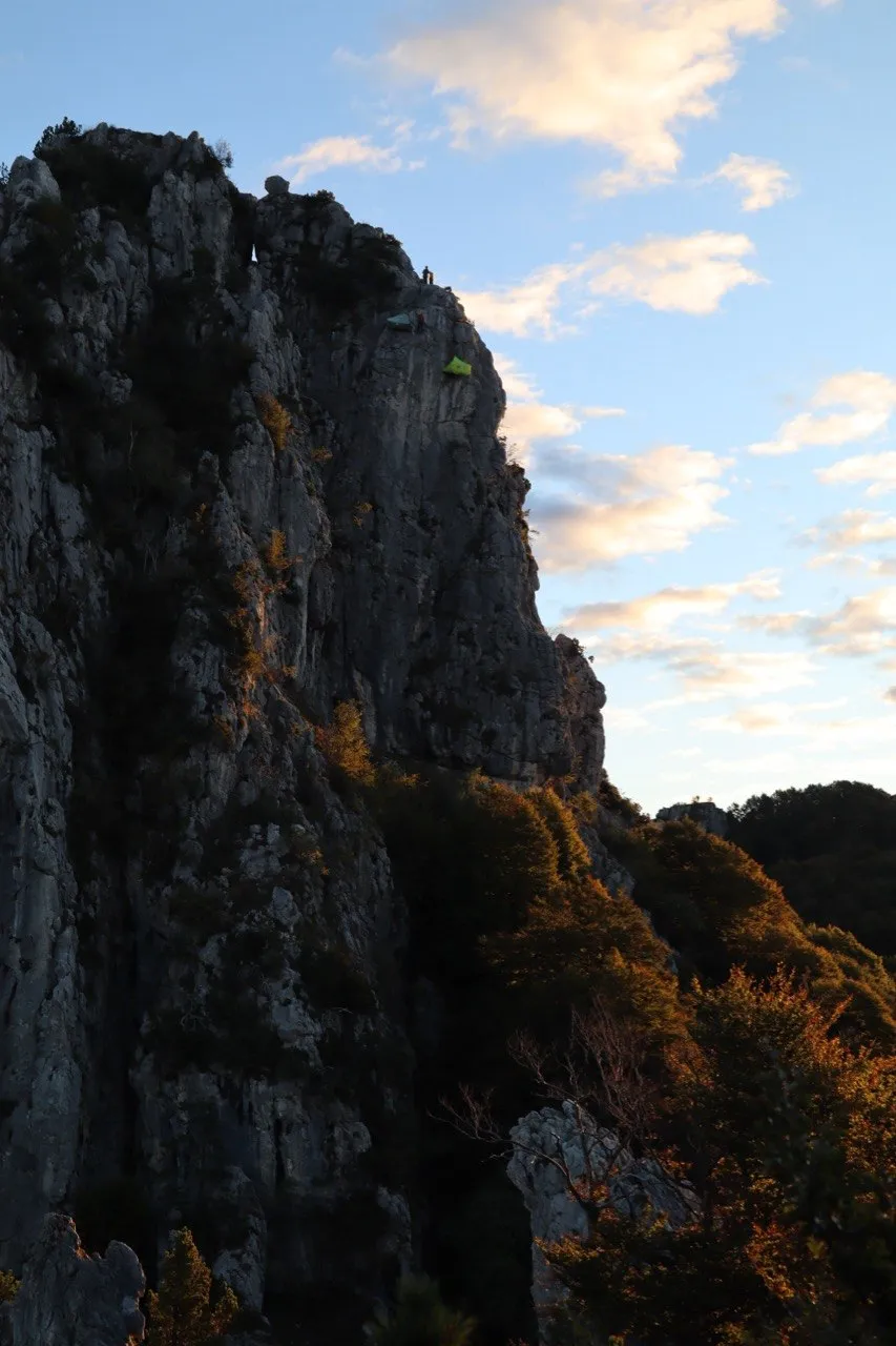 Green bivouac tent on a narrow ledge high on the limestone wall, cloudy evening sky.