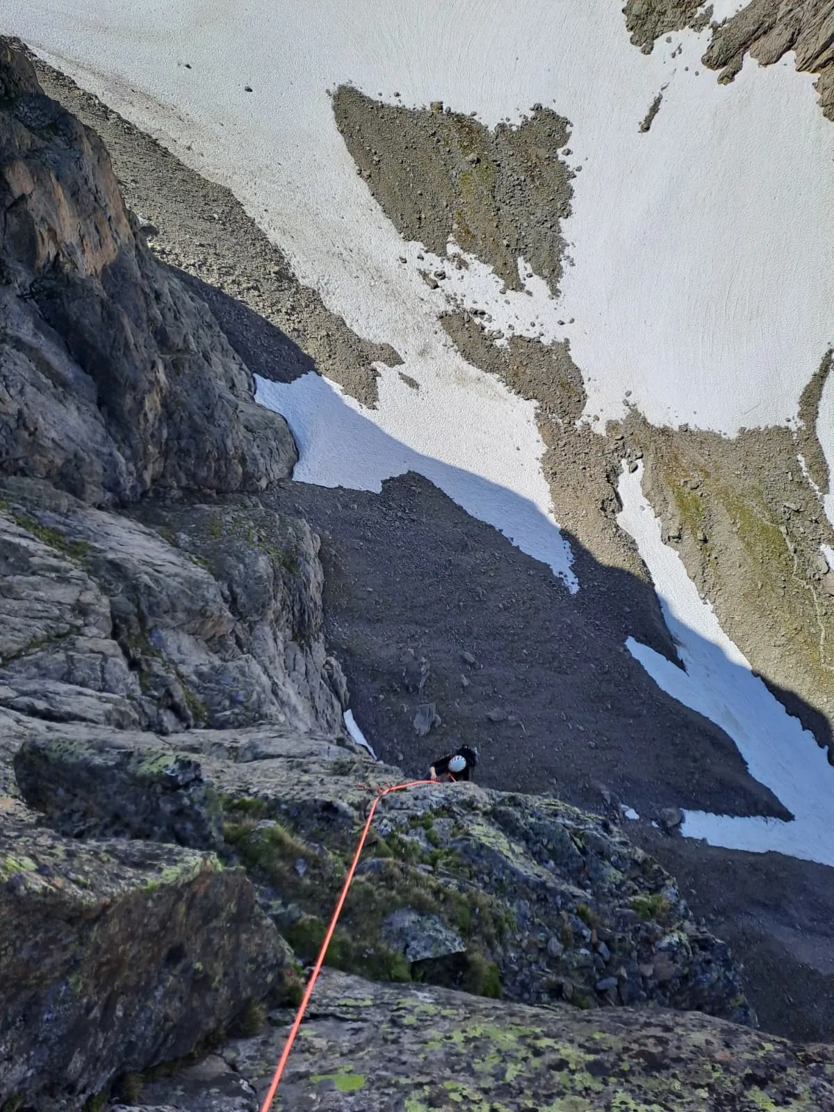 Looking down the wall at the second climbing up in a white helmet, scree slopes and old snowfields far below.
