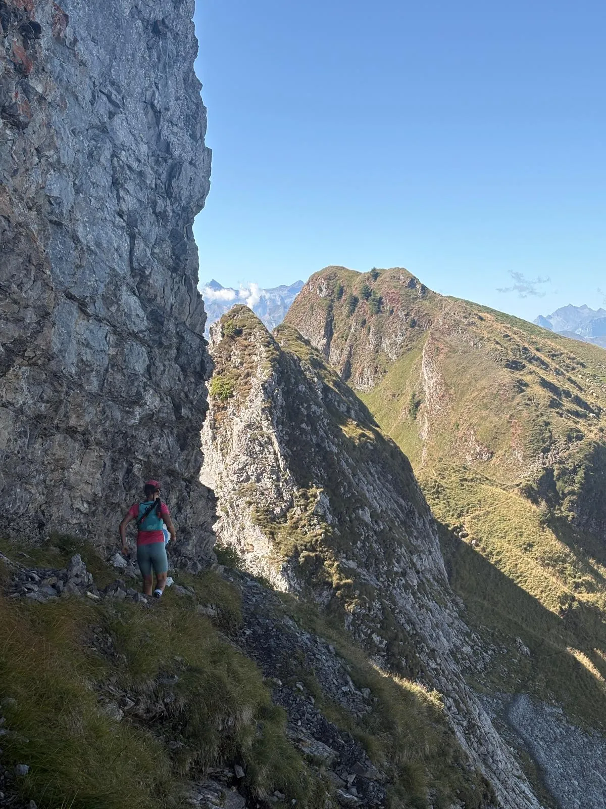 A runner traverses a narrow path beneath a big rock wall, the jagged ridge running on ahead.