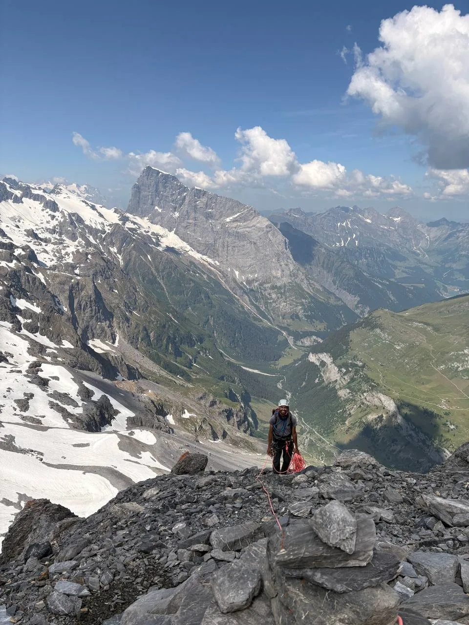 A mountaineer at a stance on broken rock, with a wide panorama of green valley, snowfields and peaks behind.