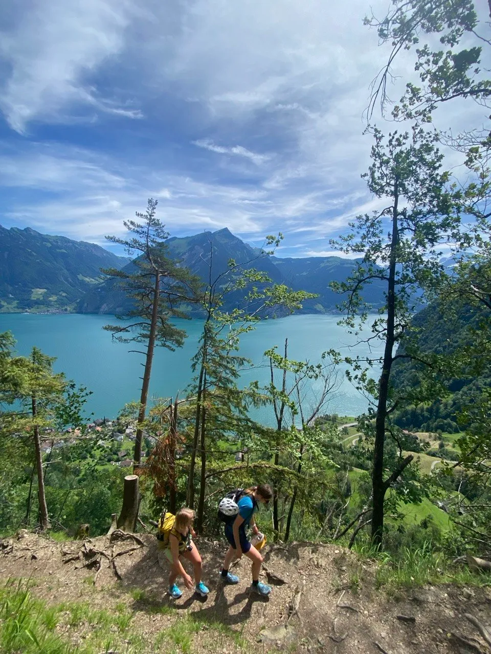 Two hikers descending a steep trail high above turquoise Lake Uri.