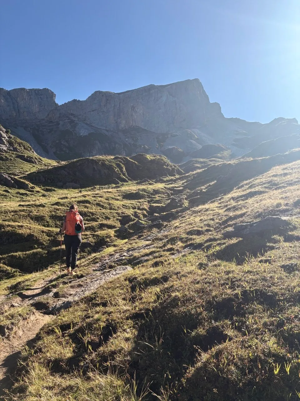 A hiker with an orange pack climbs alpine pasture toward the foot of the wall in morning light, the rocky mass of the Chaiserstock above.