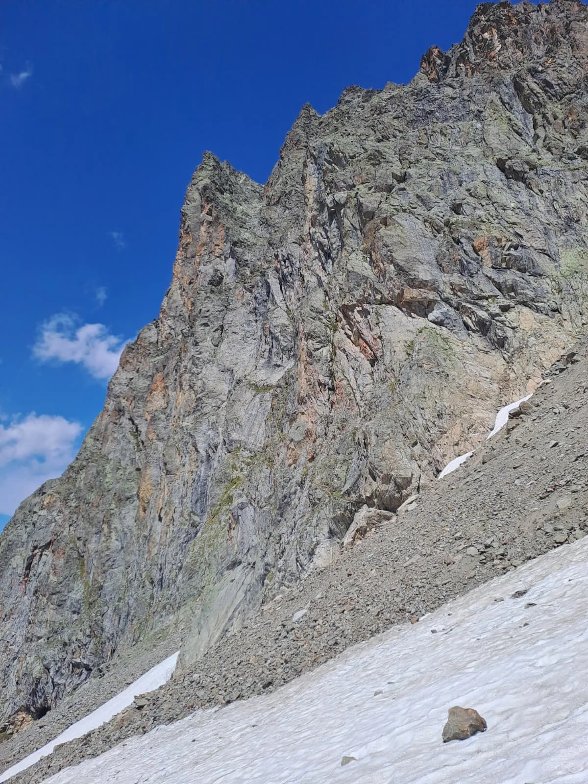The steep, rugged rock face of the Gwächten rising above scree and an old snowfield, a sharp rock spire to the left.