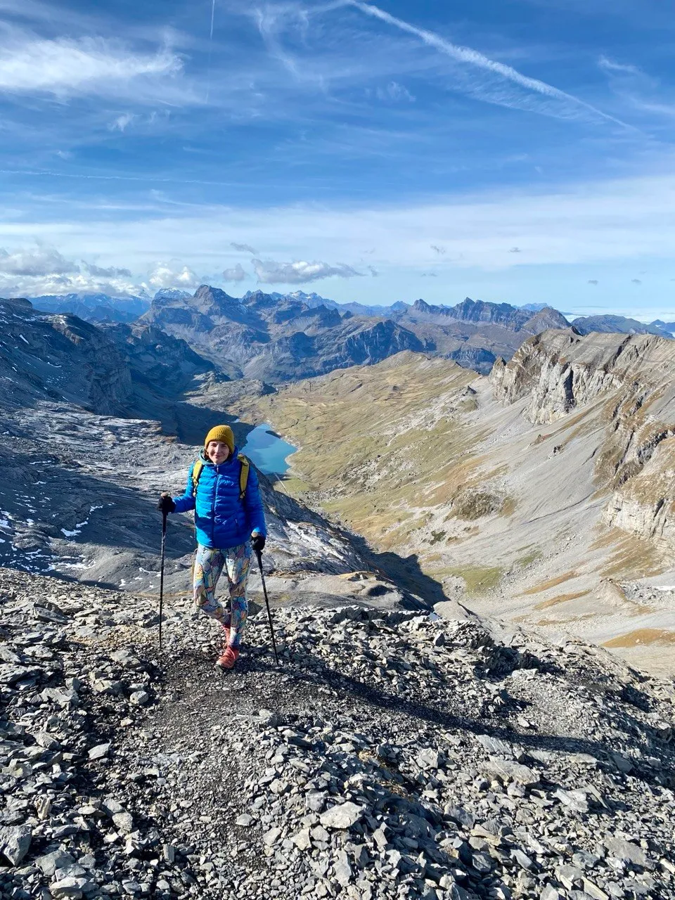 Hiker with trekking poles on the ridge, mountain lake in the background.