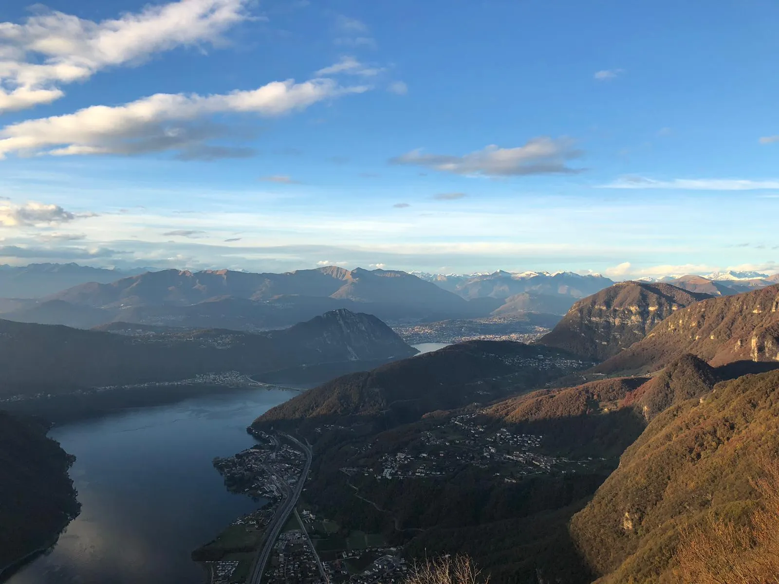 A wide panorama over Lake Lugano and hazy blue mountain ranges under a bright sky.