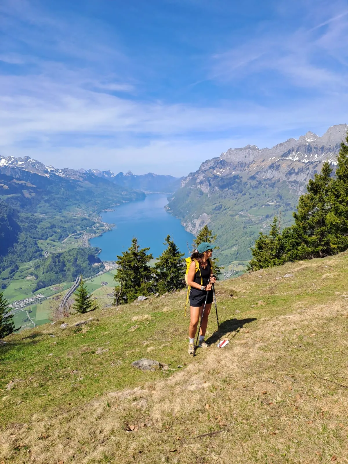 A hiker pauses on an open spring meadow with the turquoise Walensee stretching far below.