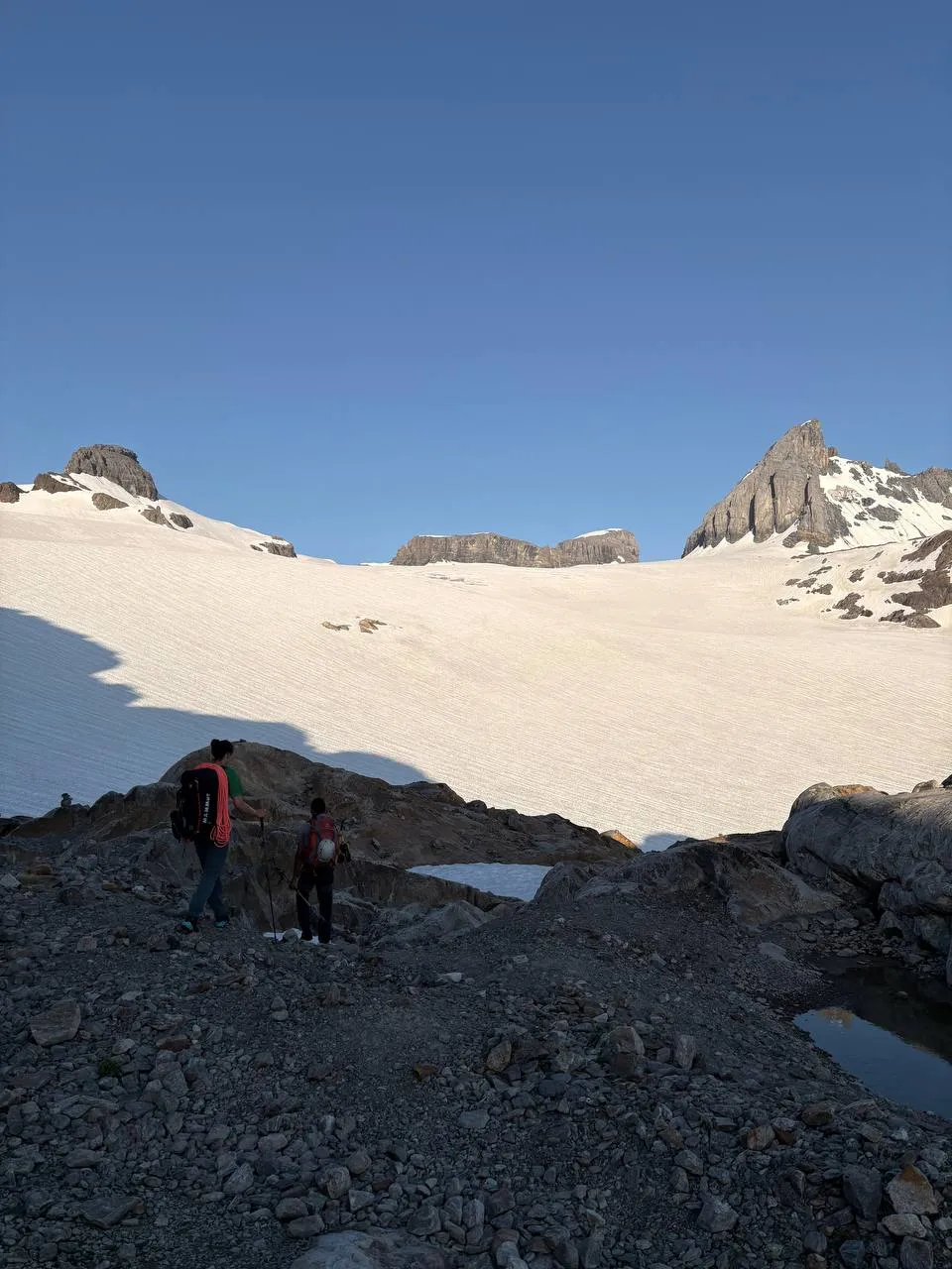 Two hikers reaching the edge of a wide snowfield at first light, rocky peaks all around.