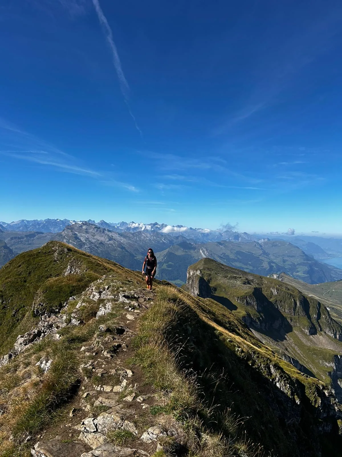 Una corredora avanza hacia la cámara por la estrecha cresta rocosa, con toda la cadena de los Alpes de Uri y una franja del lago de los Cuatro Cantones al fondo.