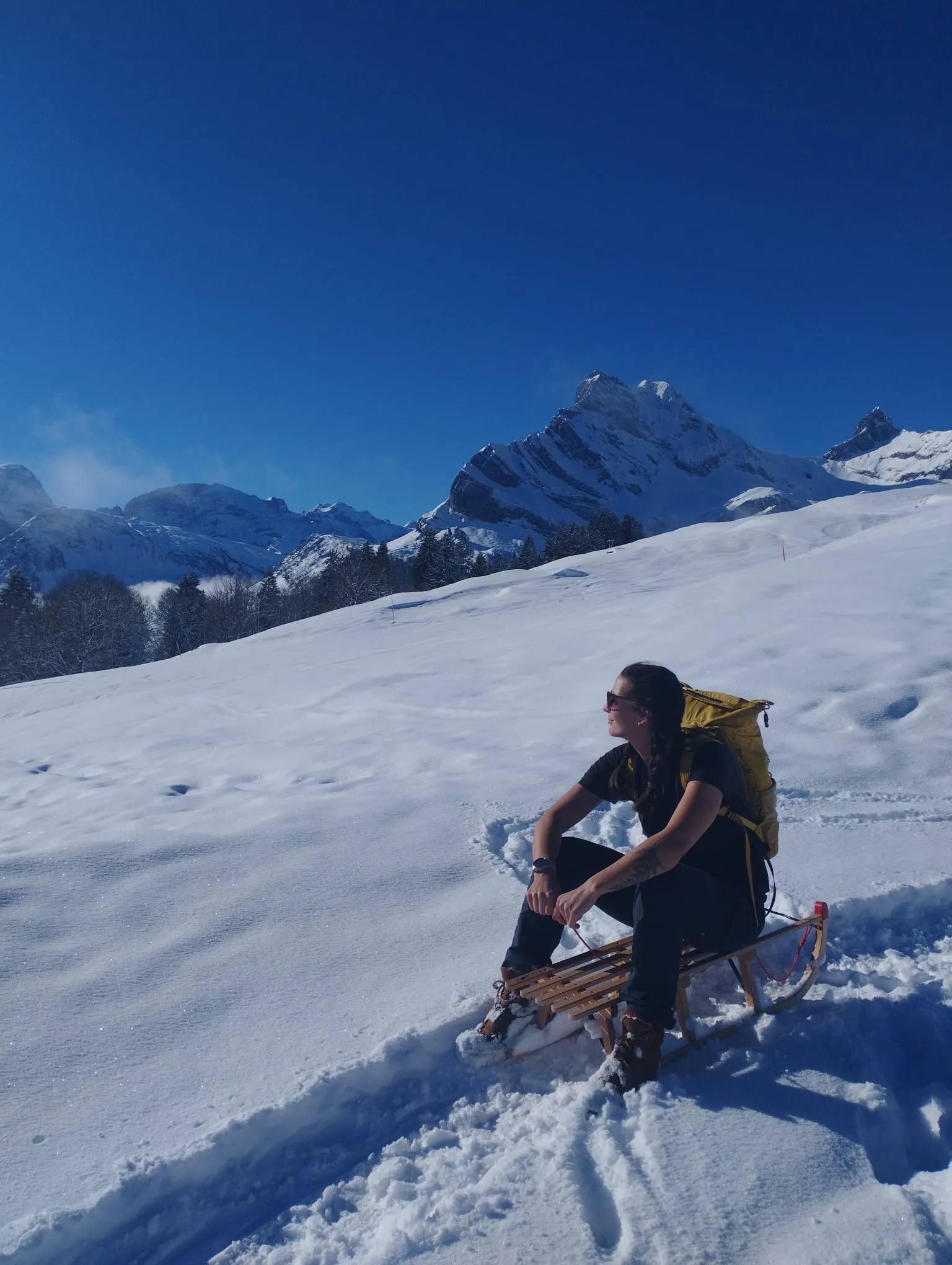 A woman sits on a wooden sledge in the snow, looking up at a striking snow-covered peak under a deep blue sky.