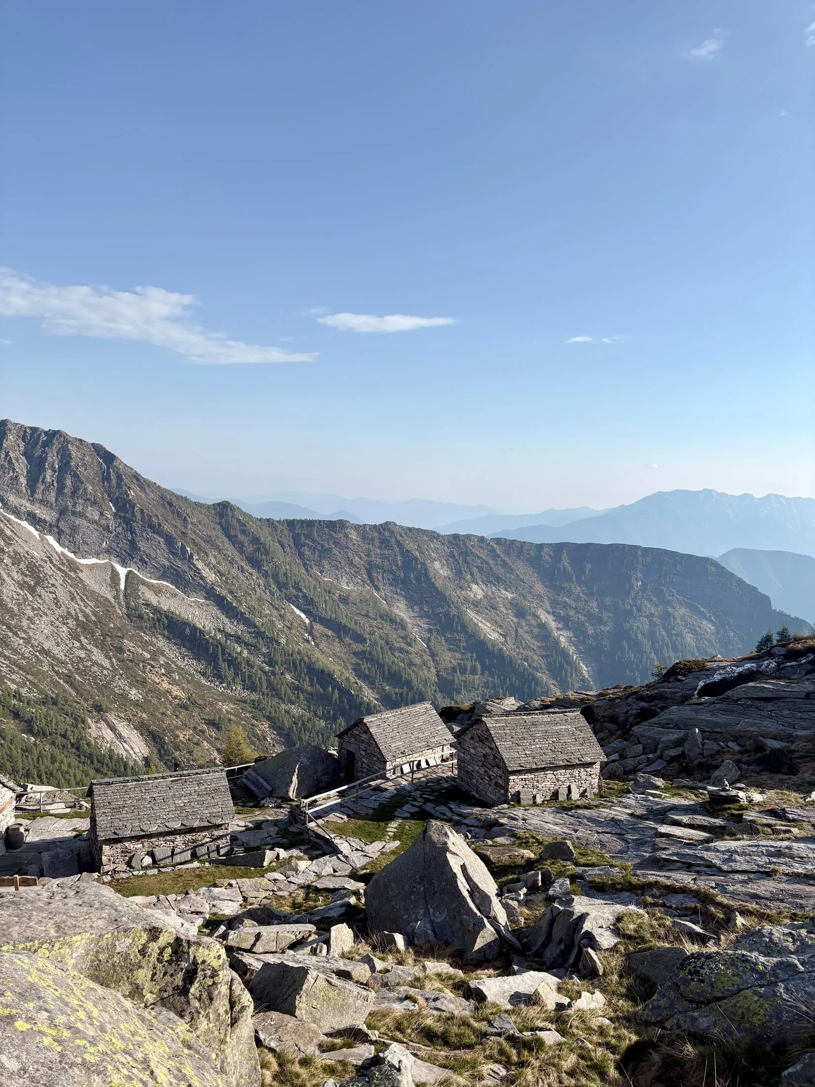 A cluster of old Ticino dry-stone huts on a rocky plateau, mountains behind.