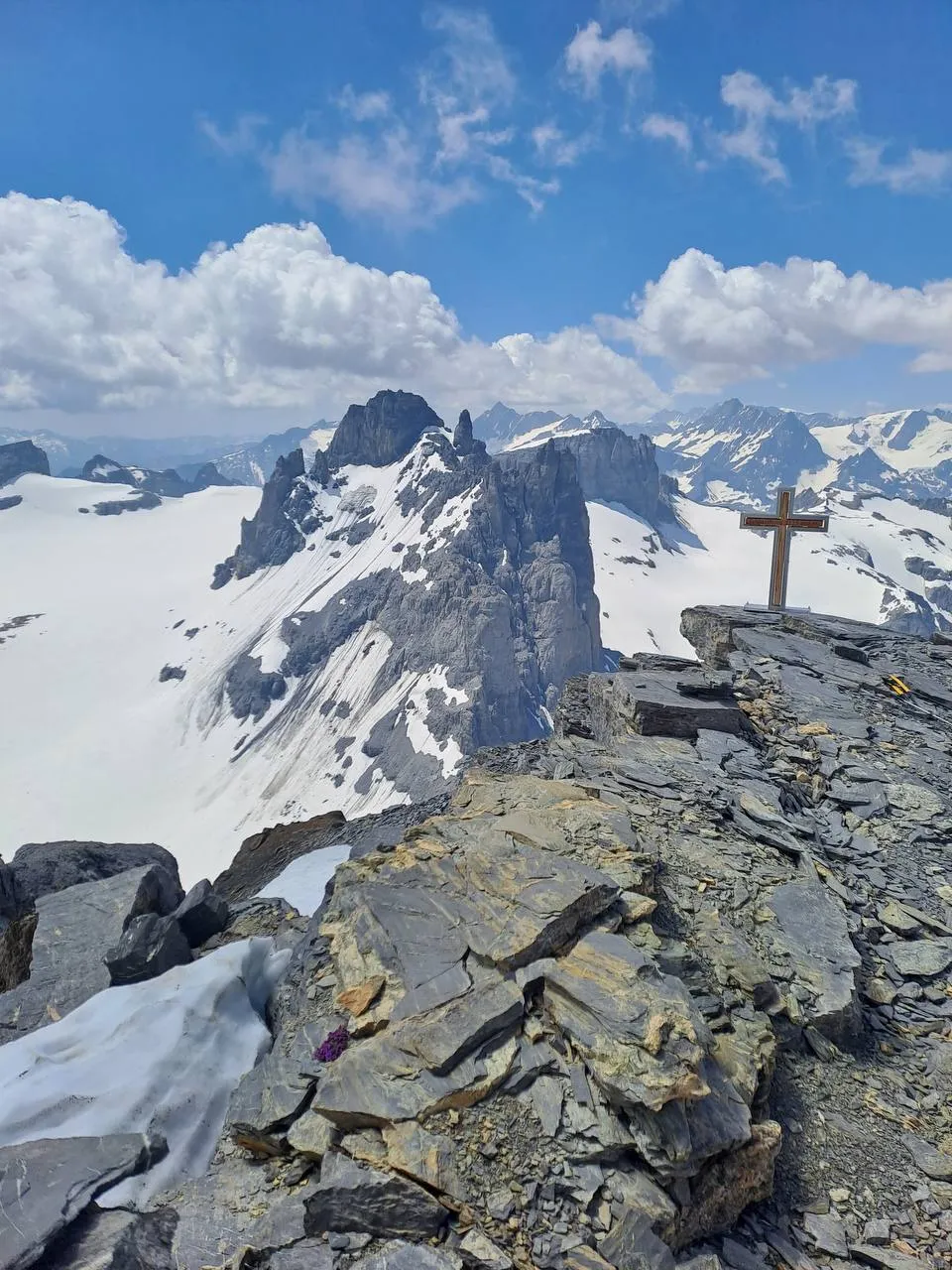 The Schlossberg summit cross on slabby rock, with snowy glaciers and jagged ridges under the clouds beyond.