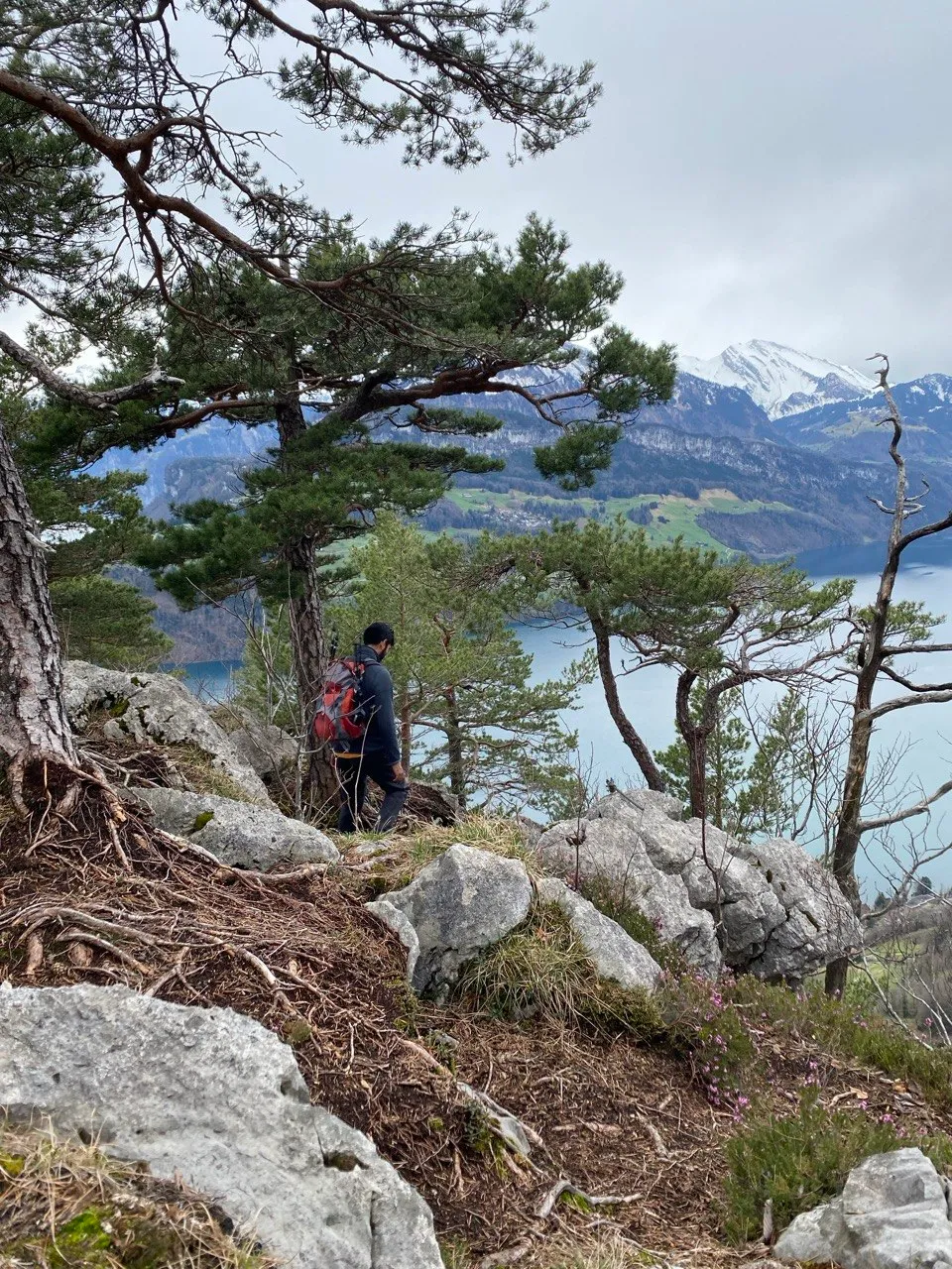 A hiker with a red backpack picks a way along the rocky, pine-studded ridge high above the lake.