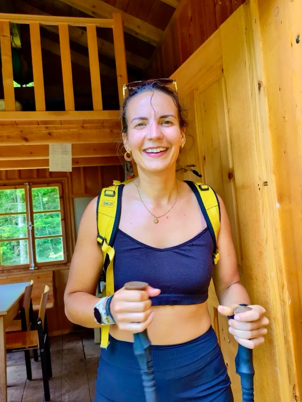 Smiling hiker with backpack and poles inside a wooden mountain hut.