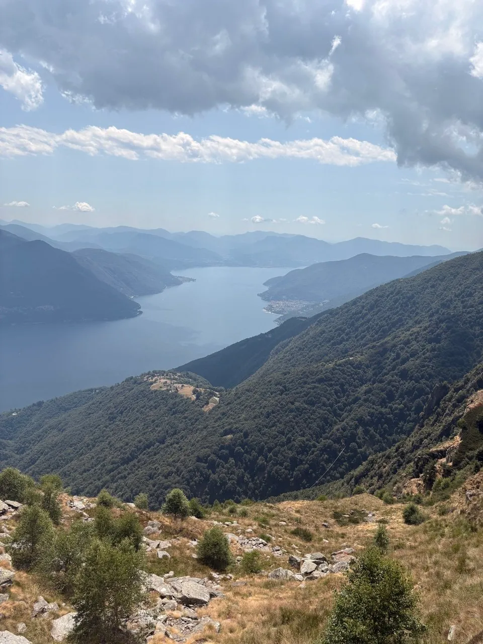 Lago Maggiore stretching south into the haze, seen from a rocky, grassy slope high above the wooded shore.