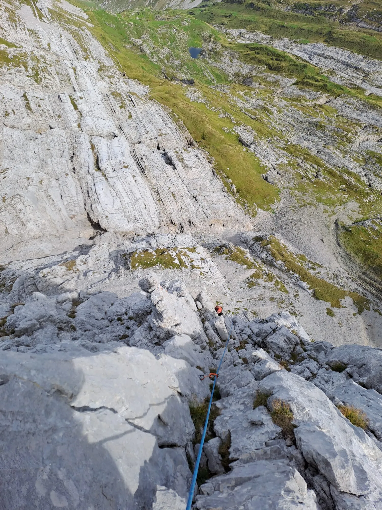 Looking straight down the wall to the belayer far below, the blue rope in the rock, a small tarn and green slopes beyond.