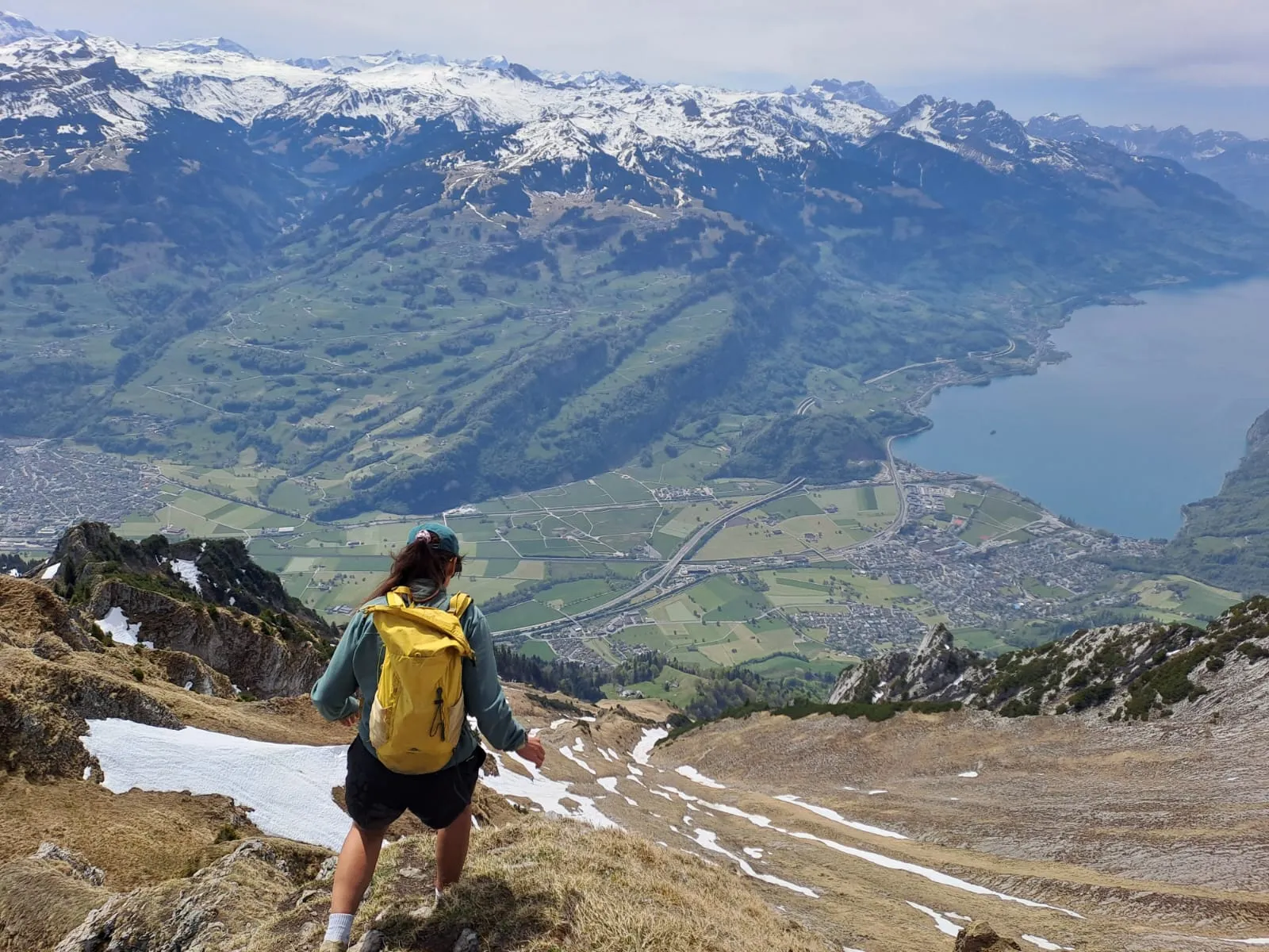 A hiker with a yellow backpack descends a grassy slope high above the Walensee, with snow-streaked Glarus peaks across the valley.