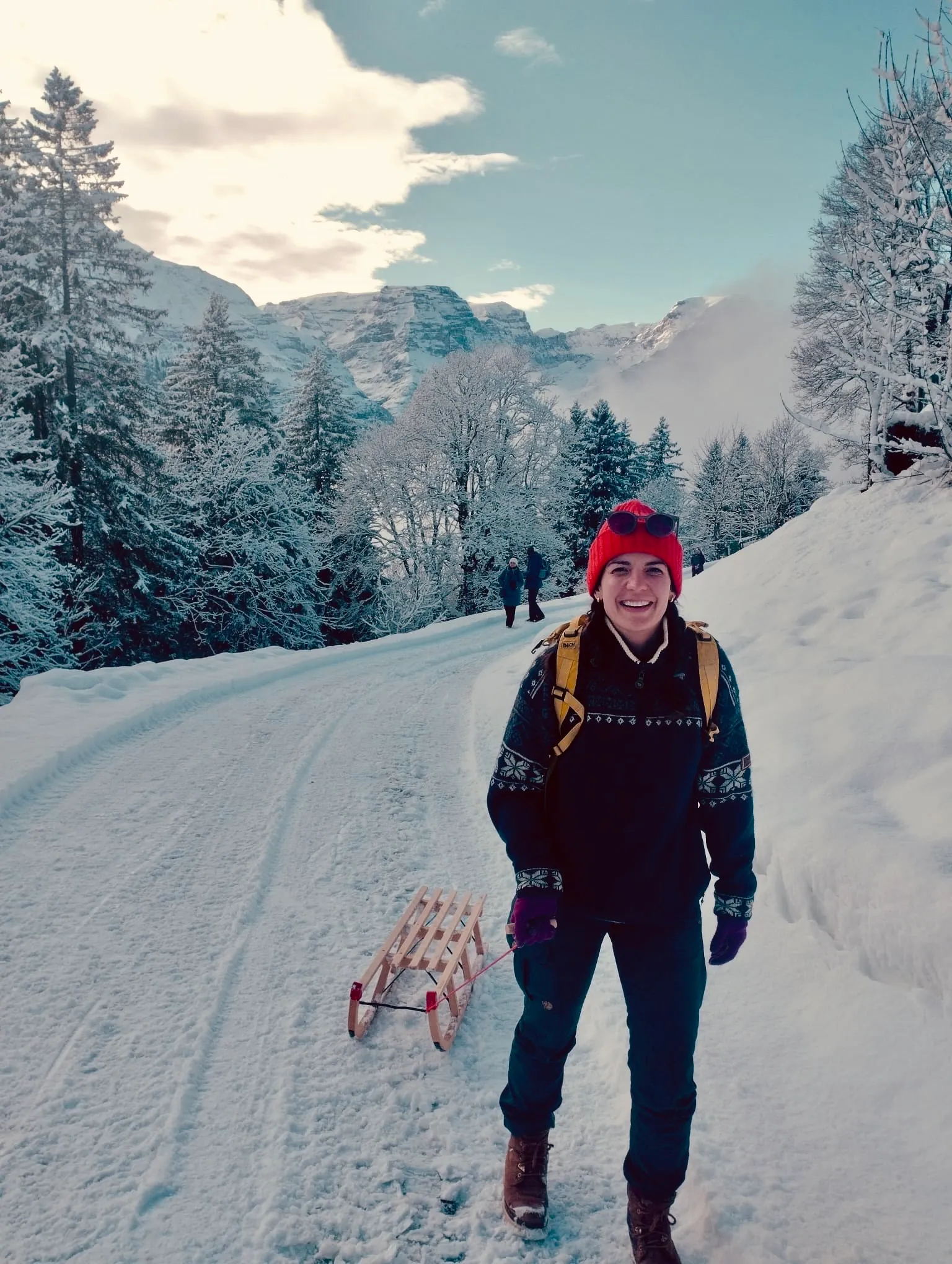 A woman in a red knit hat stands smiling next to a wooden sledge on the snowy road back to Braunwald.