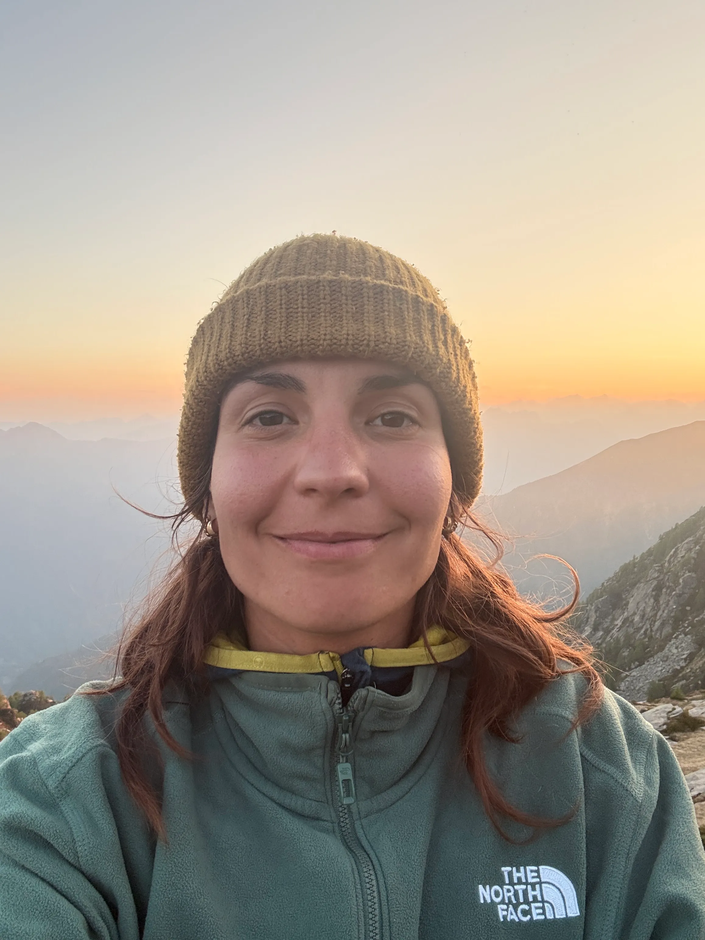 A smiling hiker in a beanie and fleece at golden hour, hazy Ticino peaks fading into the light behind her.
