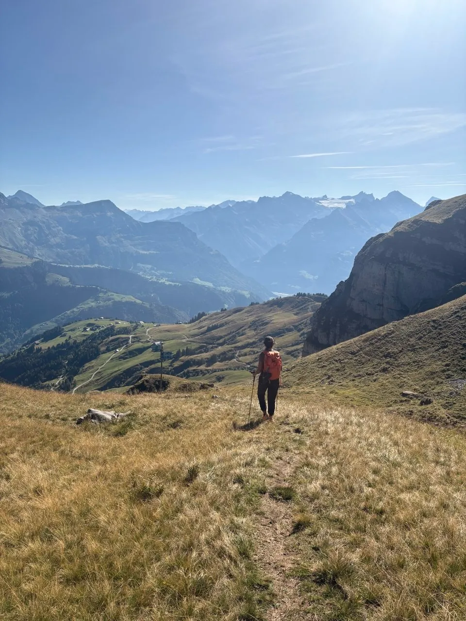 Hiker on a green grassy slope looks out across valleys to a wide chain of Alpine peaks.