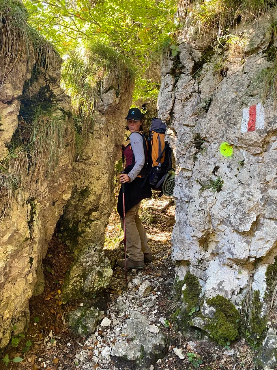Hiker squeezing through a narrow rock crevice, red-and-white trail marker on the wall.