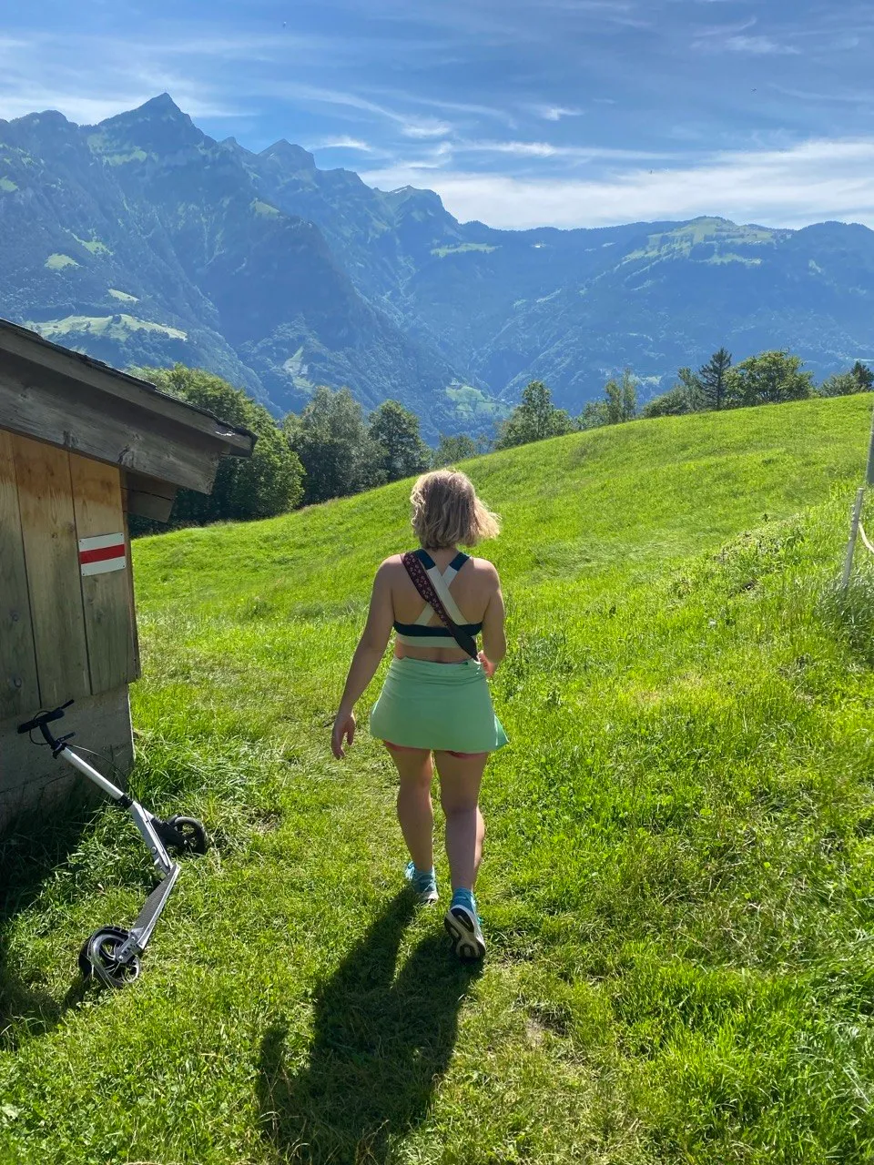 Hiker walking toward the mountains past a hut, kick-scooter parked nearby.
